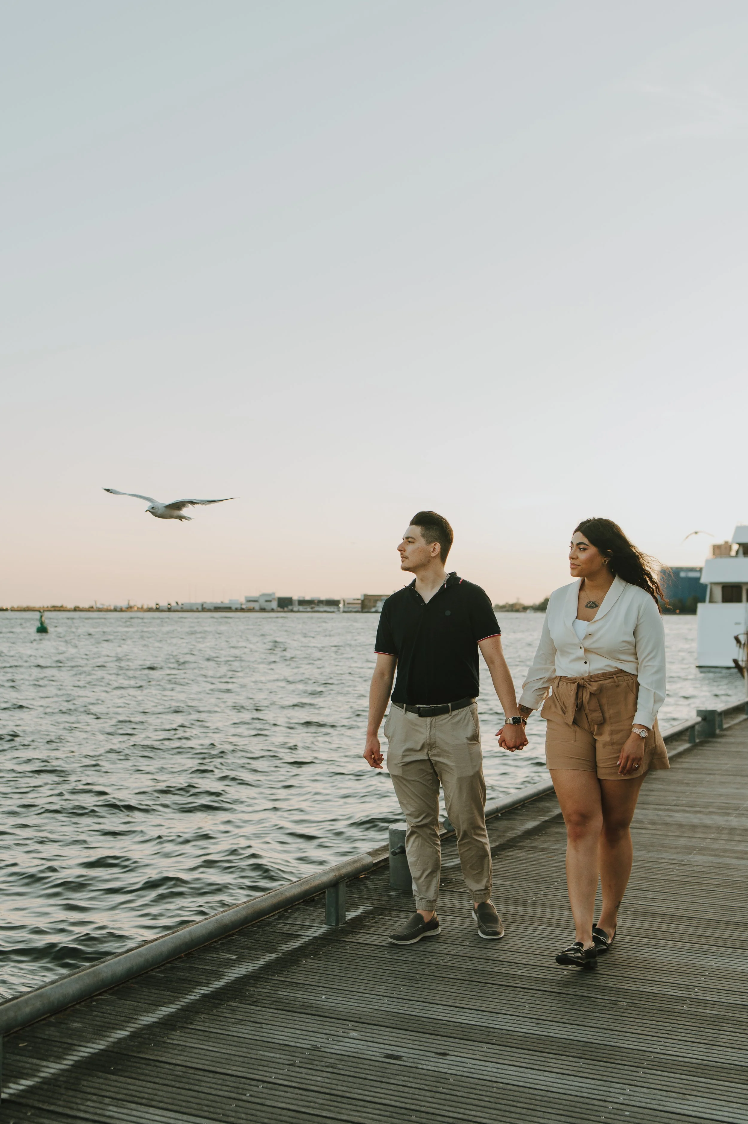 A couple walking hand-in-hand along a waterfront pier during sunset, with a seagull flying nearby and boats in the background.