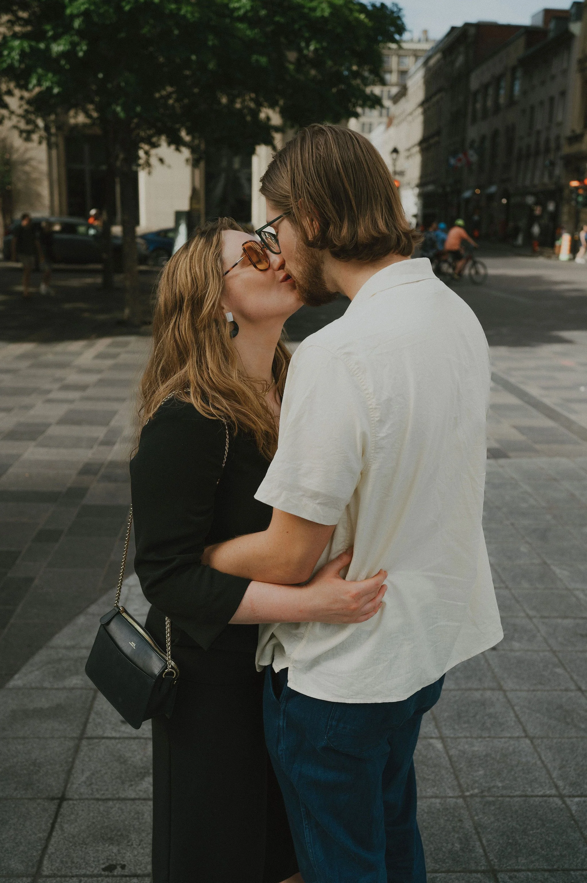 A couple kissing on a city street, with the woman wearing sunglasses and carrying a small purse.