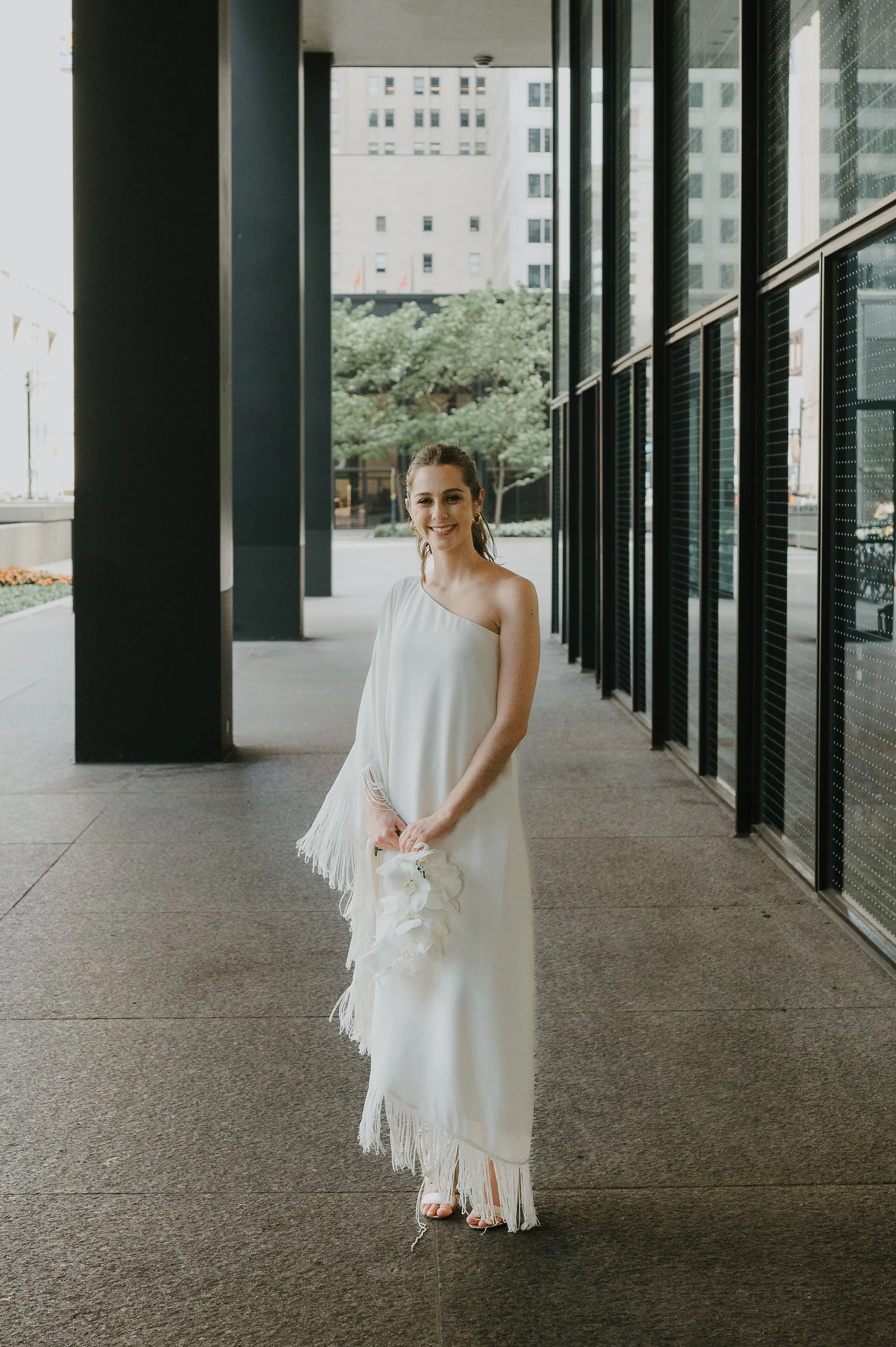 A woman in a white dress standing outside in an urban setting with tall buildings and glass windows.