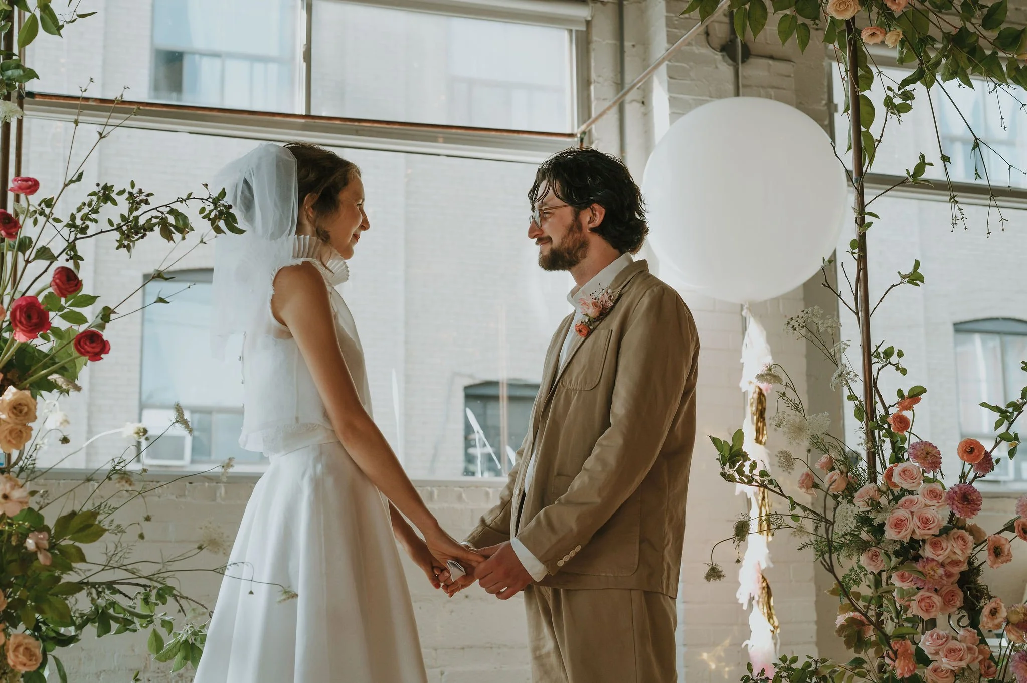 A bride and groom holding hands, facing each other, during a wedding ceremony inside a bright room decorated with flowers and a large white balloon.