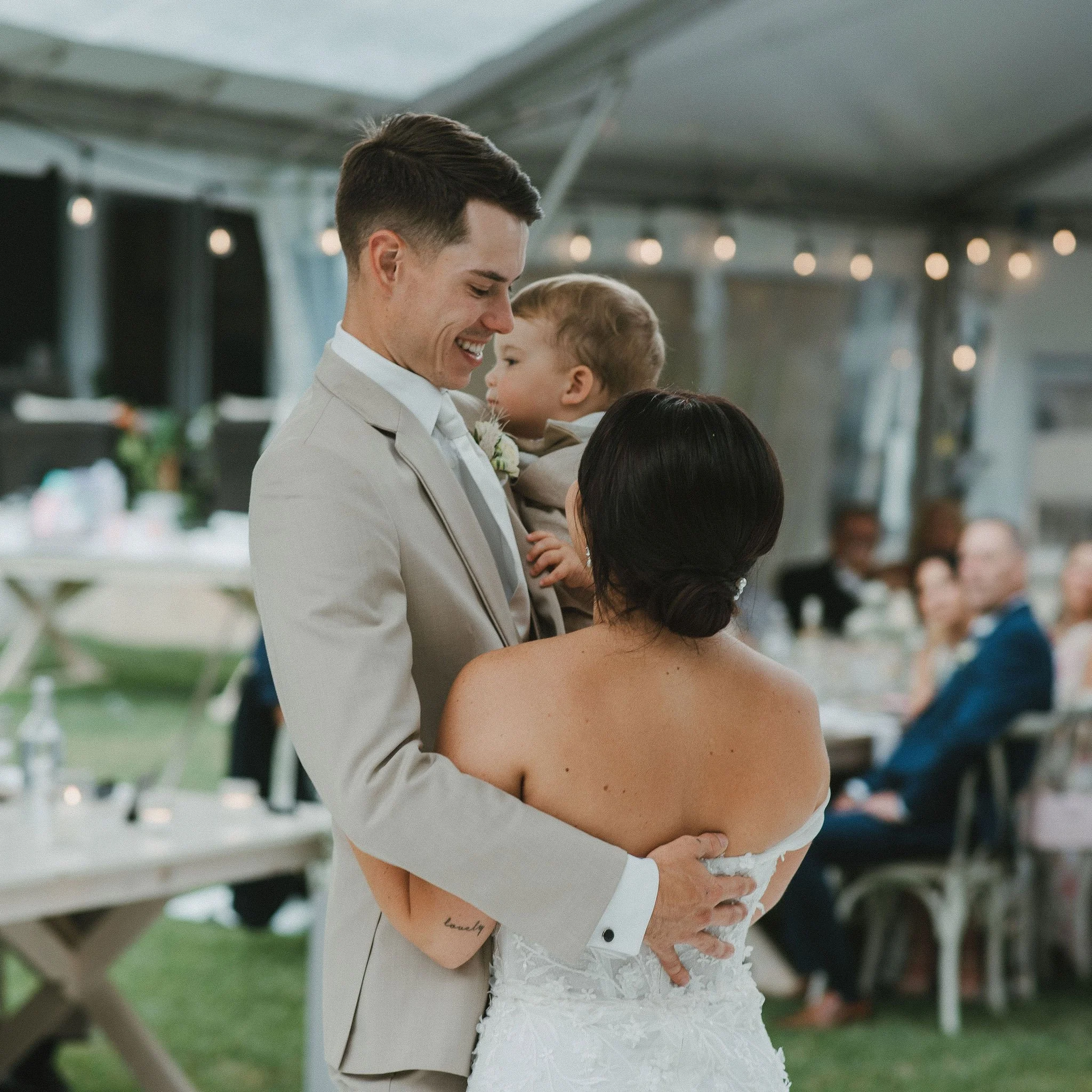A bride and groom sharing a moment at their wedding reception, holding each other while the groom smiles at the bride, who is facing away. The groom is holding a young child in his arms, and guests are seated at tables in the background under a tent.