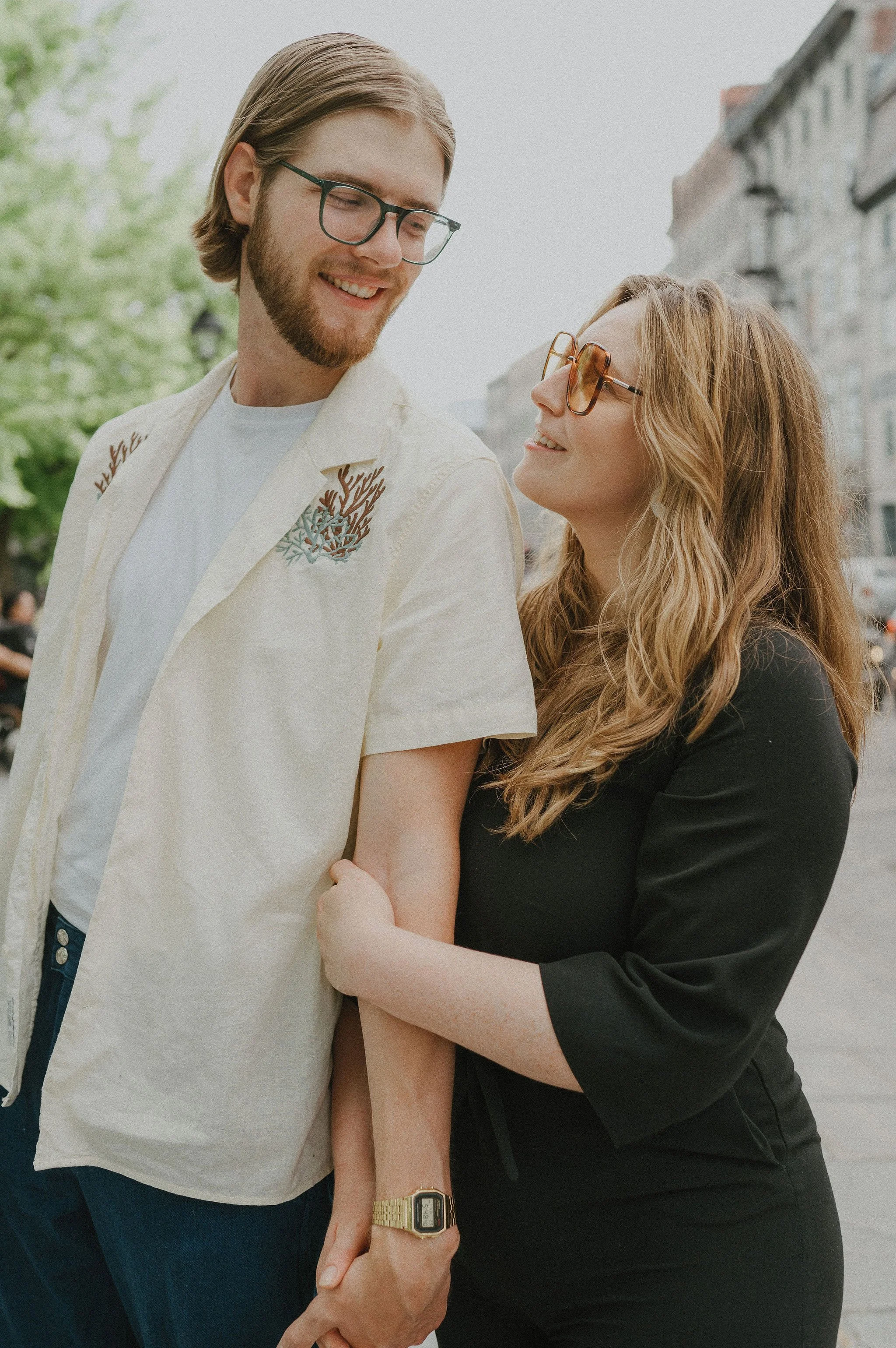 A young man and woman smiling and holding hands on a city street.