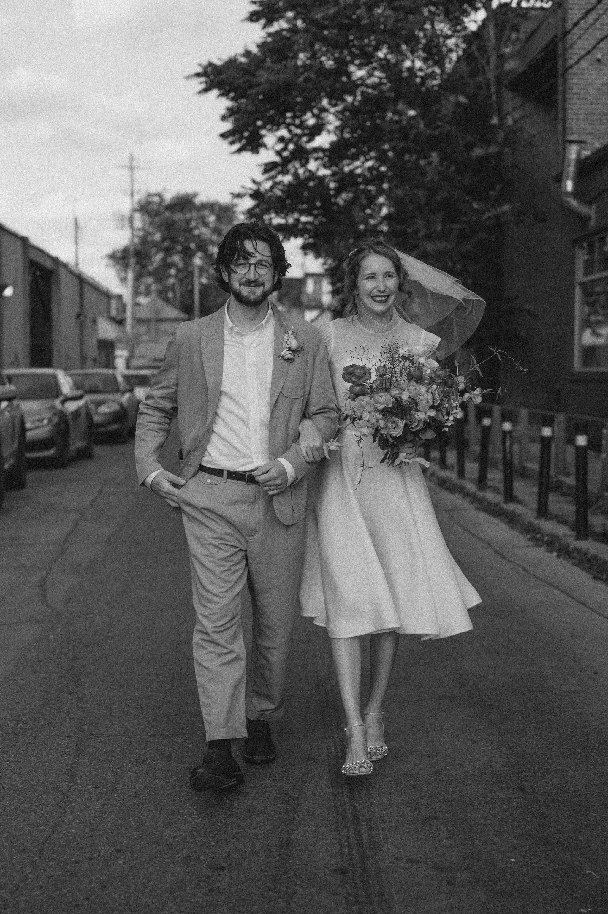 A black-and-white photo of a smiling bride and groom walking arm-in-arm on a city street. The bride is holding a large bouquet of flowers and wearing a vintage-style dress and veil. The groom is dressed in a light-colored suit with glasses. Cars and 