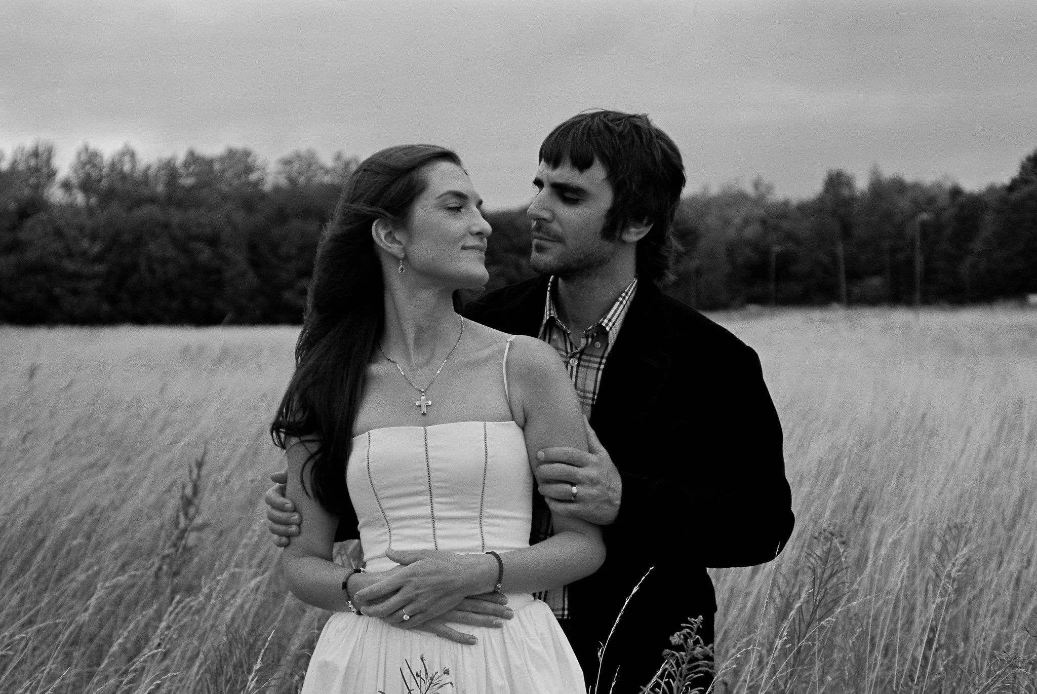 A black and white photo of a couple standing in a field of tall grass. The woman is wearing a strapless dress with jewelry, and the man is in a dark jacket and plaid shirt. They are holding each other and looking into each other's eyes.