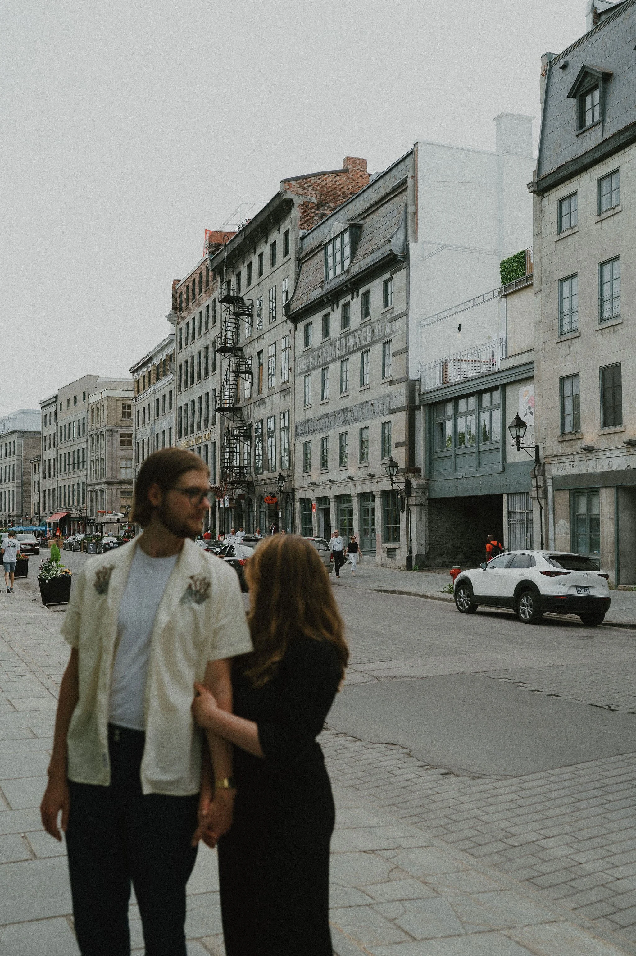 A man and woman holding hands on a city sidewalk with buildings and parked cars in the background.