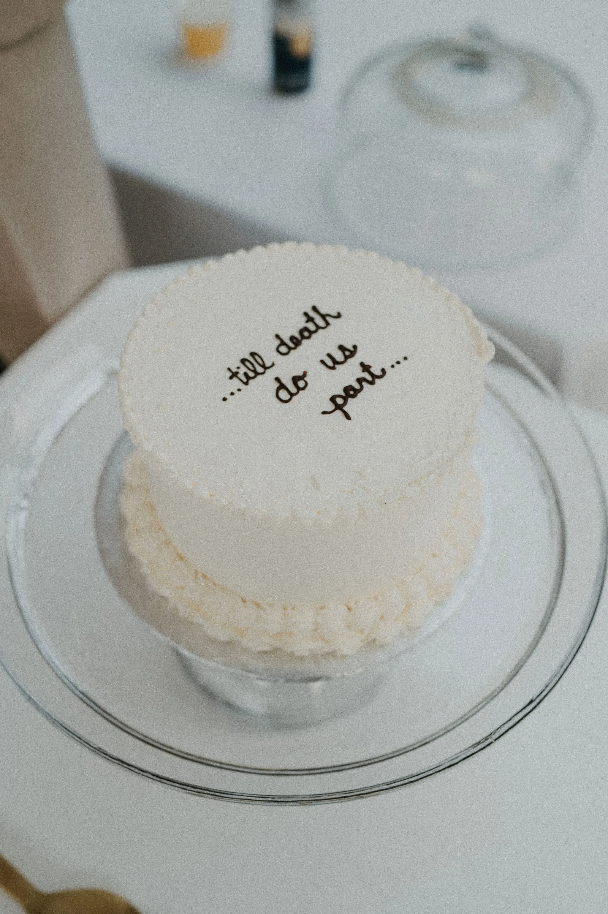A white cake with the message '..till death do us part...' written on top in black, decorative writing, sitting on a clear glass cake stand.