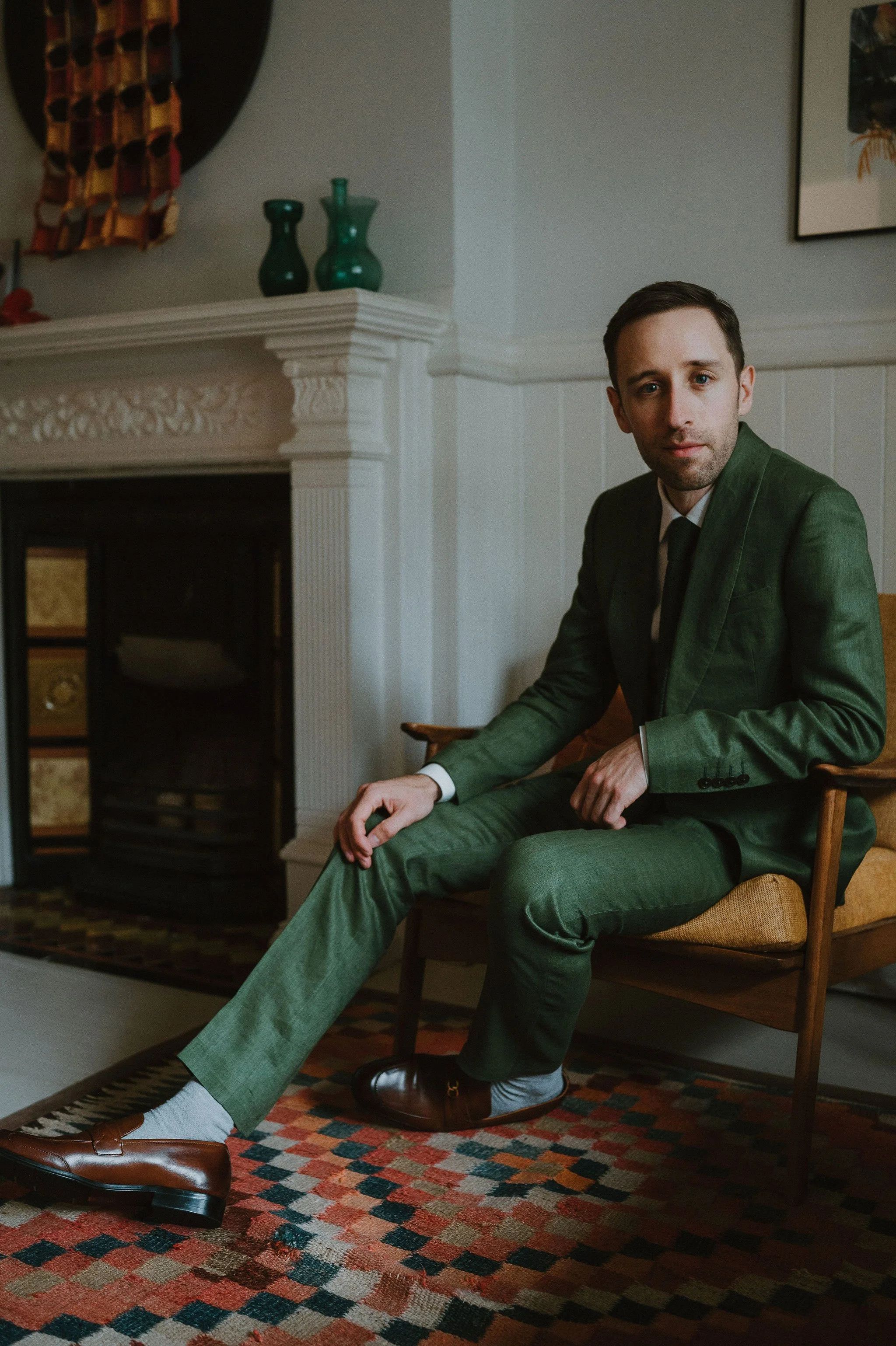 A man in a green suit sitting on a chair in a room with a fireplace and colorful decor.