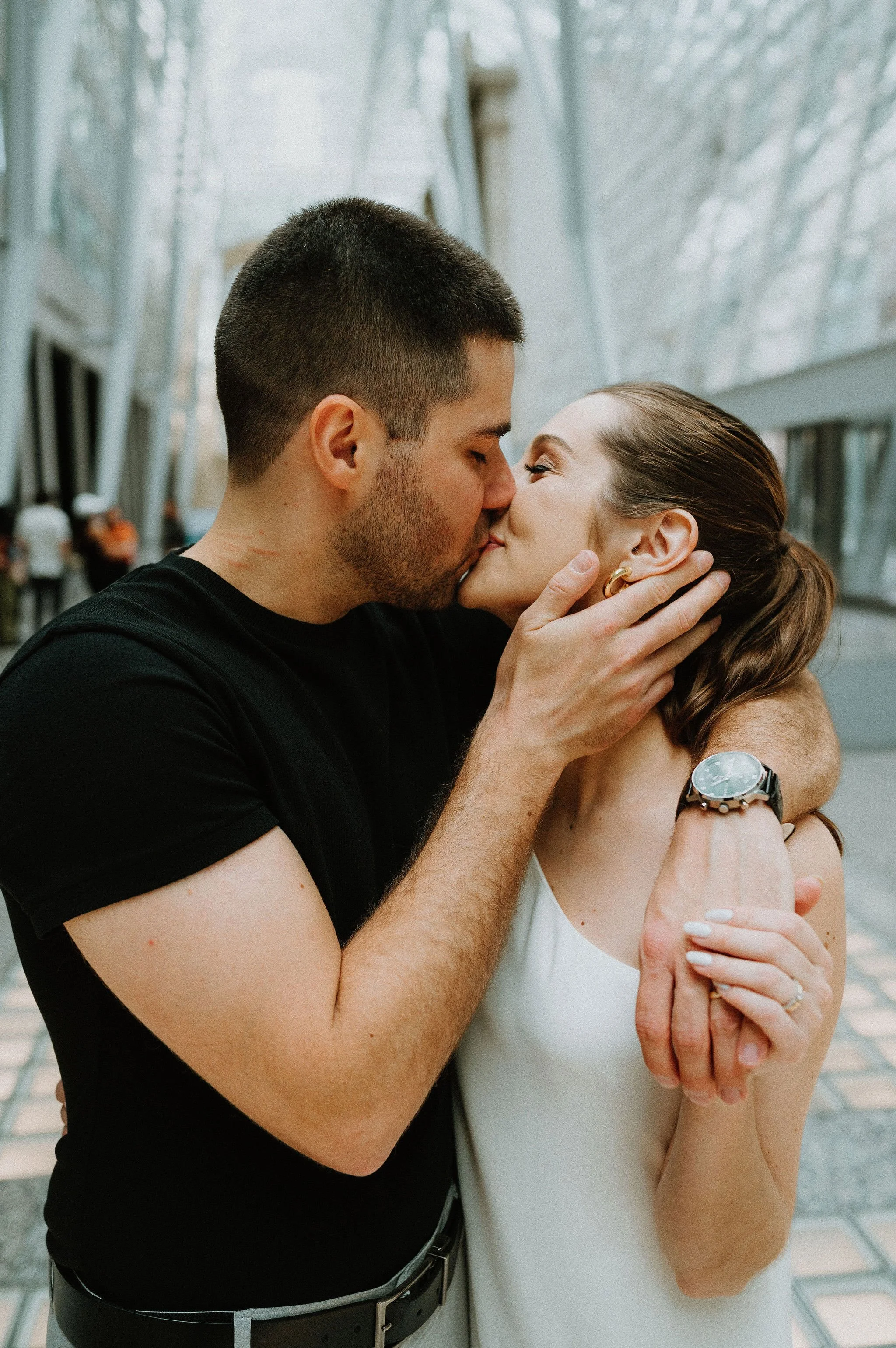A romantic couple kissing inside a modern glass building, with the man holding the woman's face gently.