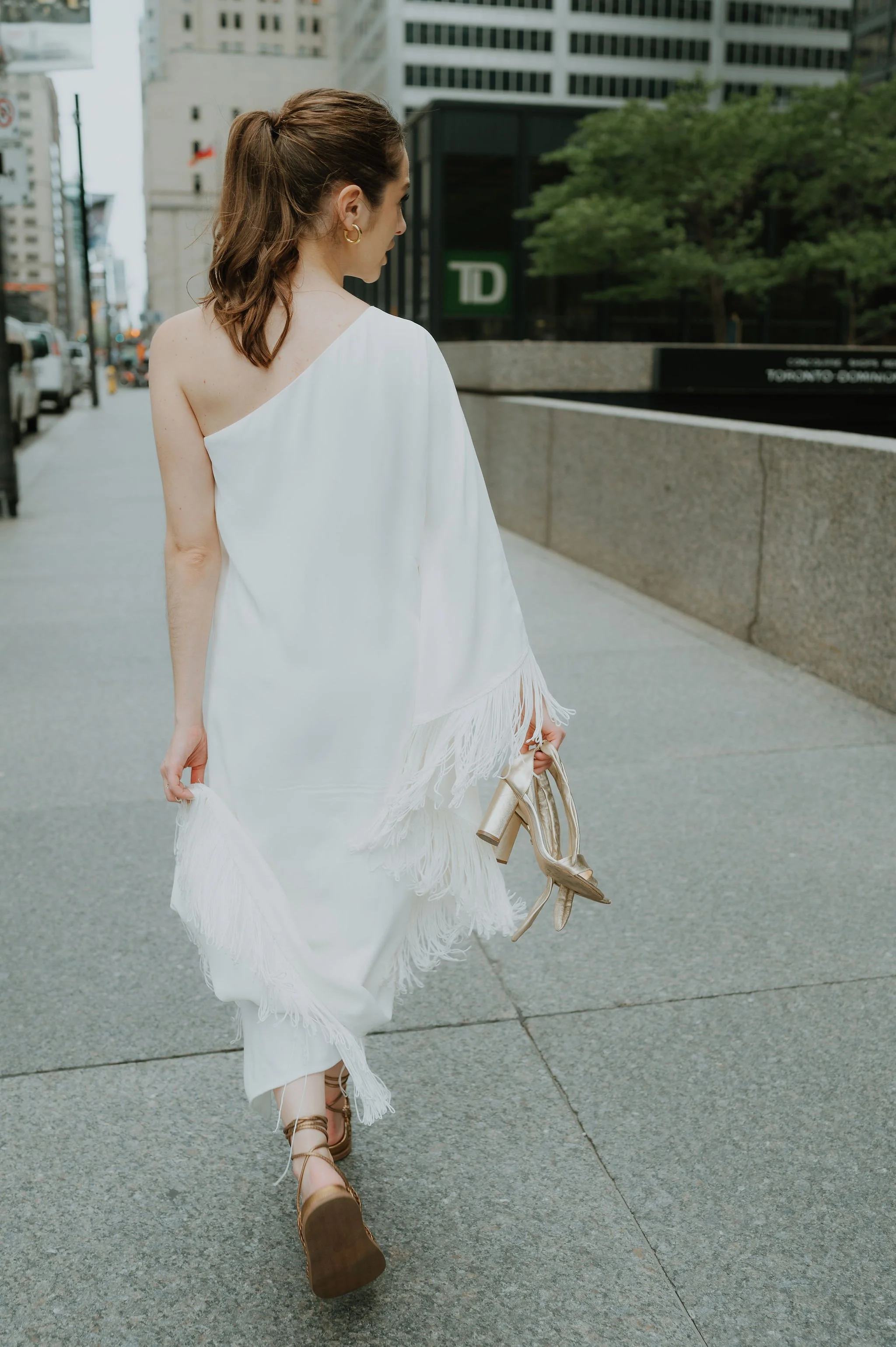 A woman wearing a white fringed dress is walking down a city sidewalk, holding a pair of high-heeled shoes in her hand.