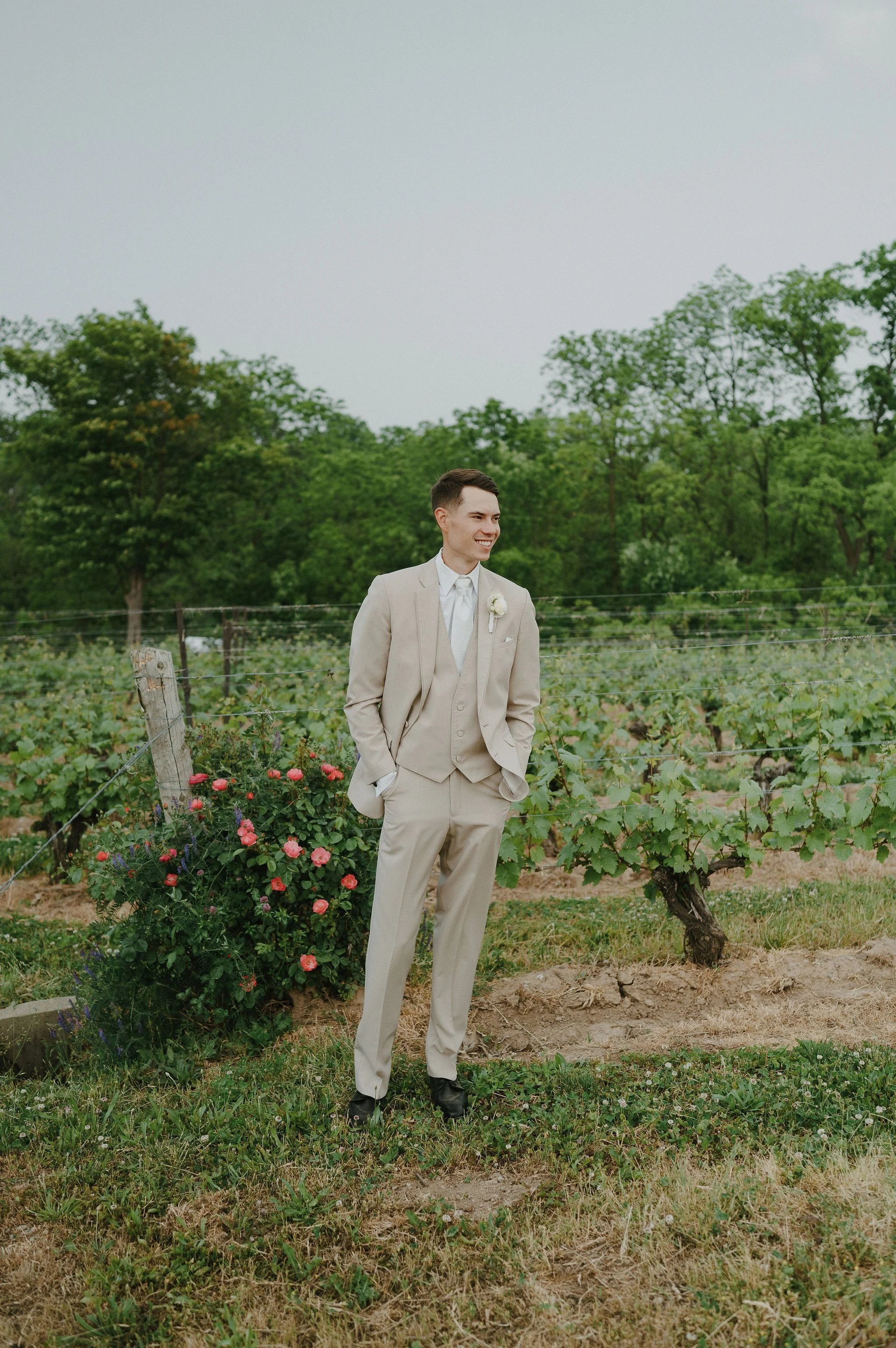 A young man in a beige suit and white shirt standing outdoors in front of a green vineyard with flowering bushes, smiling.