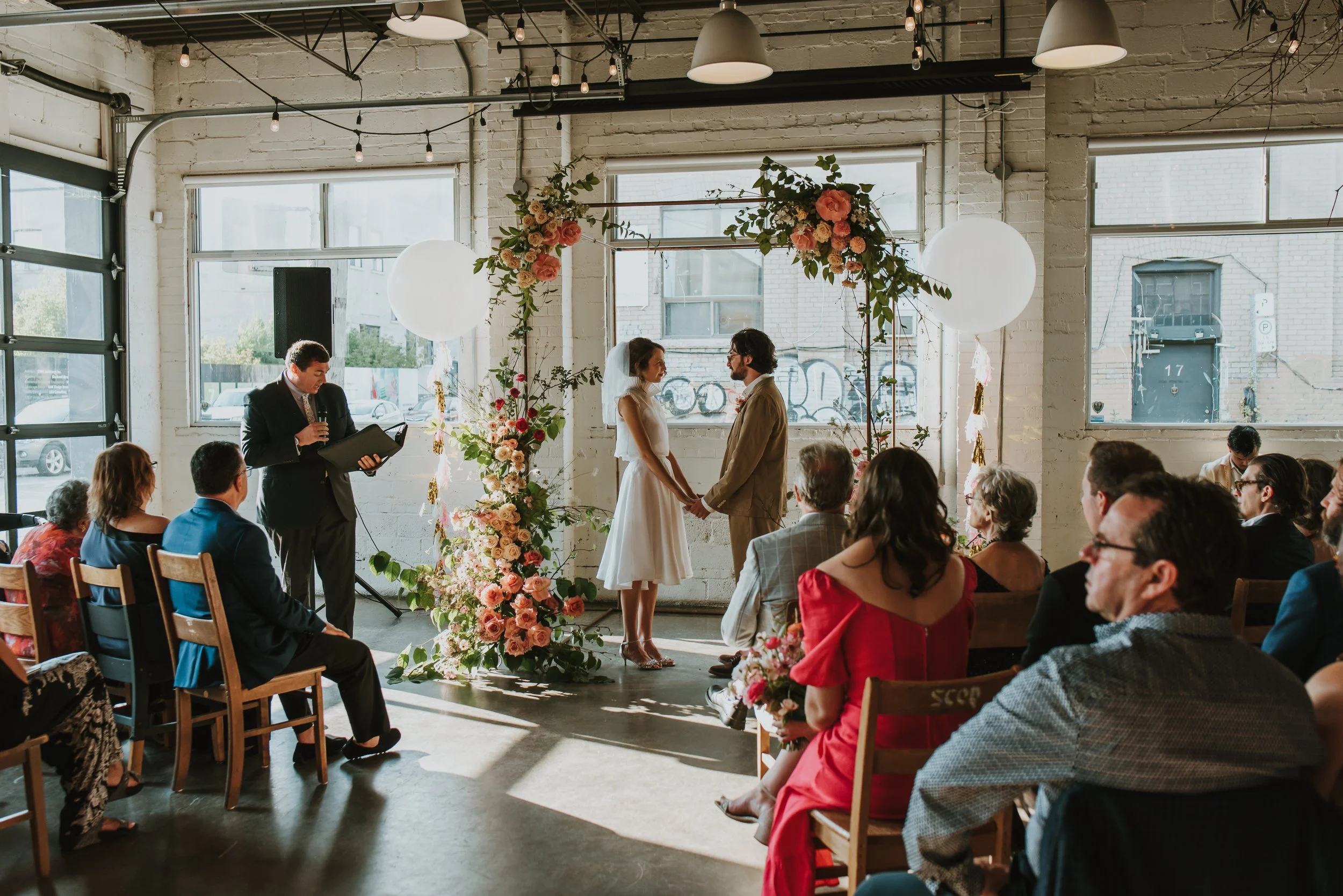A wedding ceremony taking place in a bright industrial-style space with large windows, floral decorations, and an officiant reading from a book. The bride and groom are holding hands and facing each other, surrounded by seated guests.