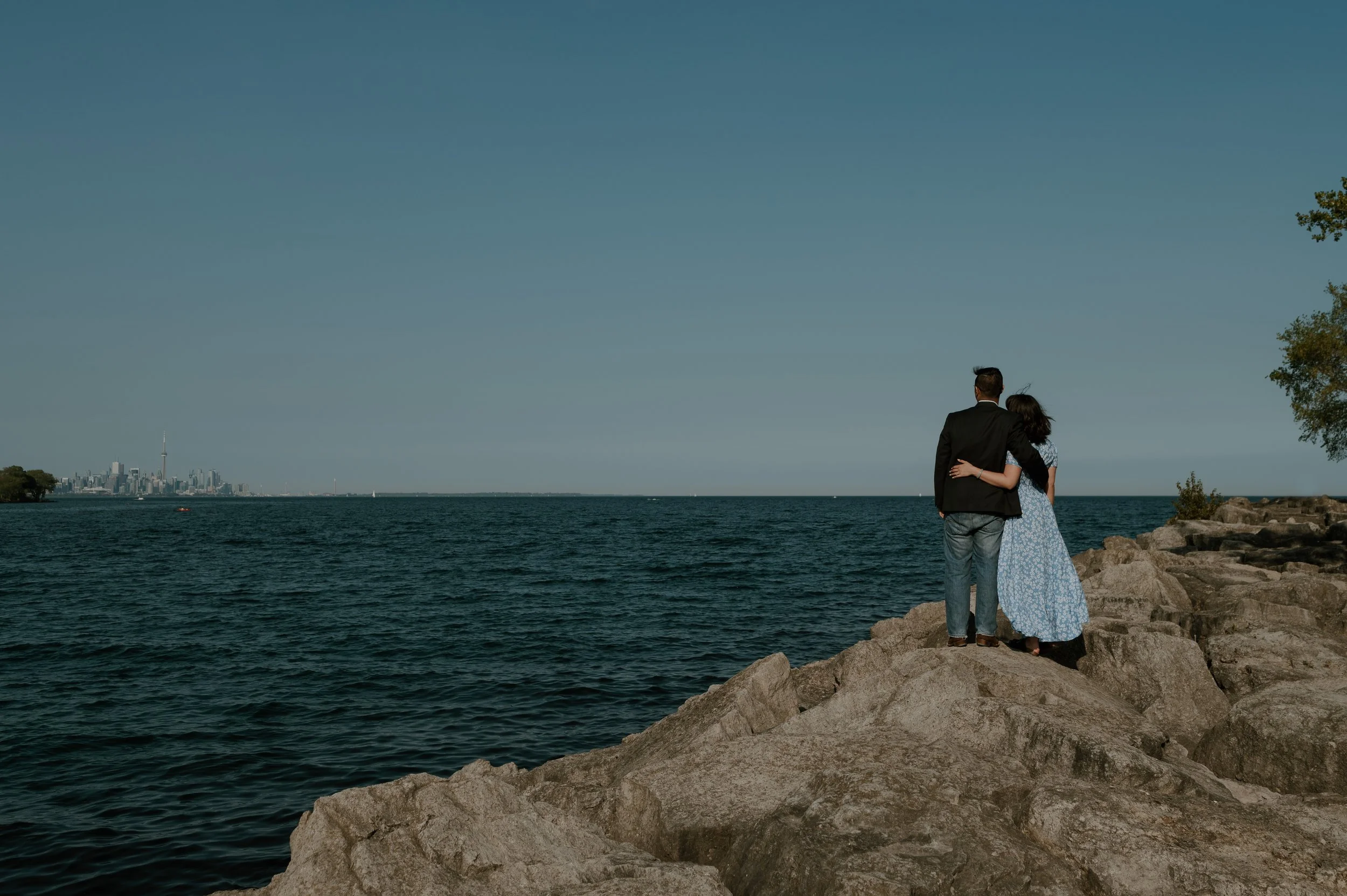 A couple stands on rocks by a large body of water, with a city skyline in the distance and a tree on the right.