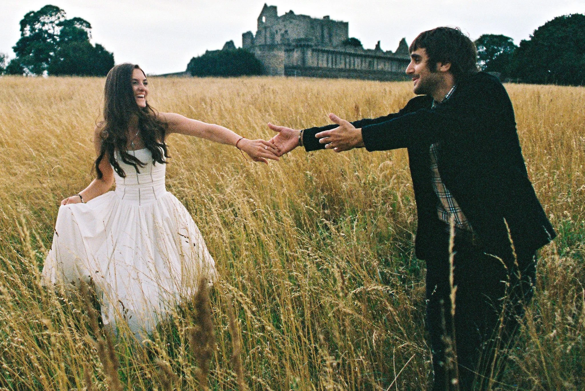 A couple is in a field of tall grass, with a castle in the background. The woman, wearing a white dress, is reaching out and holding the man's hand. The man, dressed in dark clothing, is smiling and extending his hand toward her, as if they are about