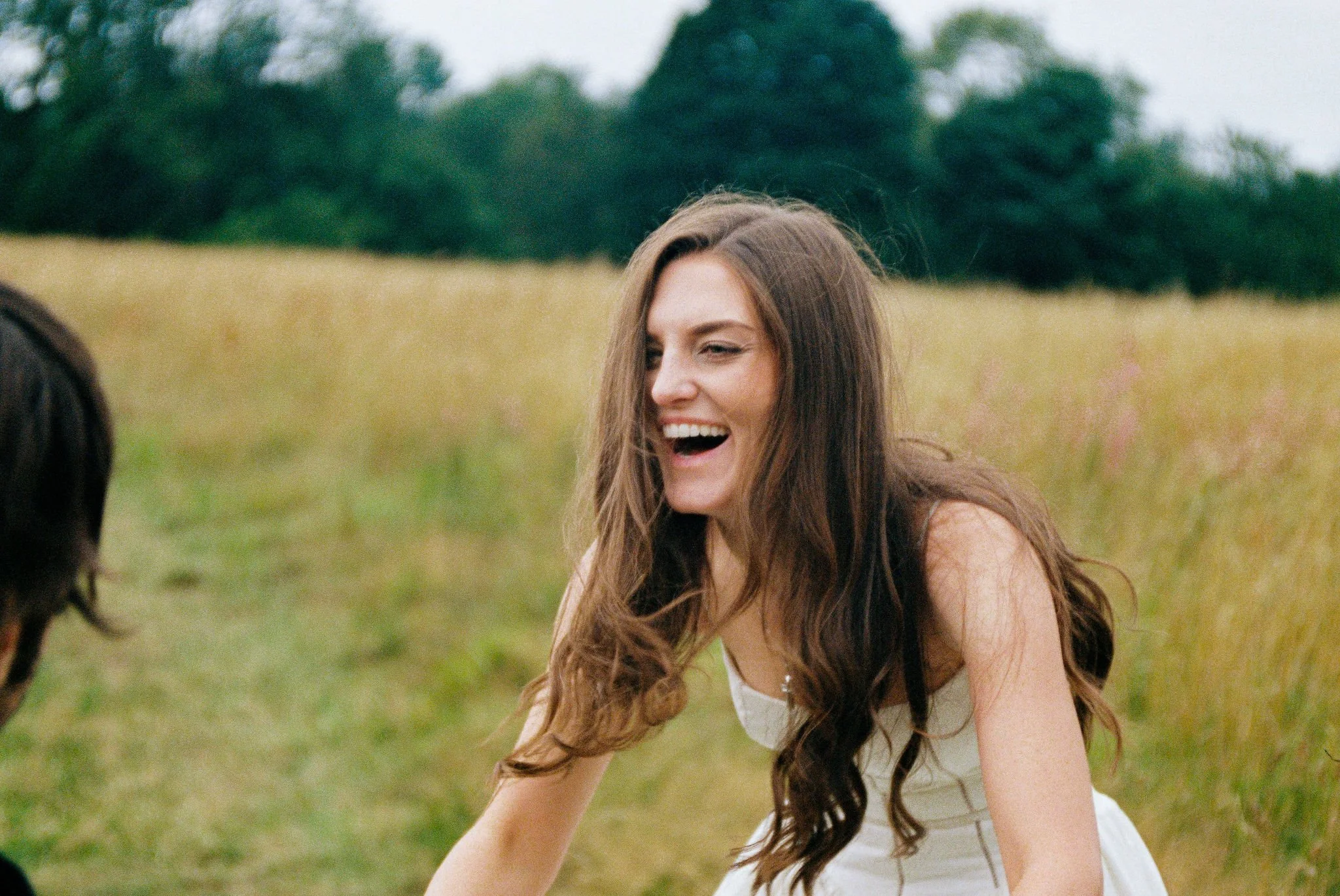A young woman with long, wavy brown hair is laughing outdoors in a grassy field with trees in the background.