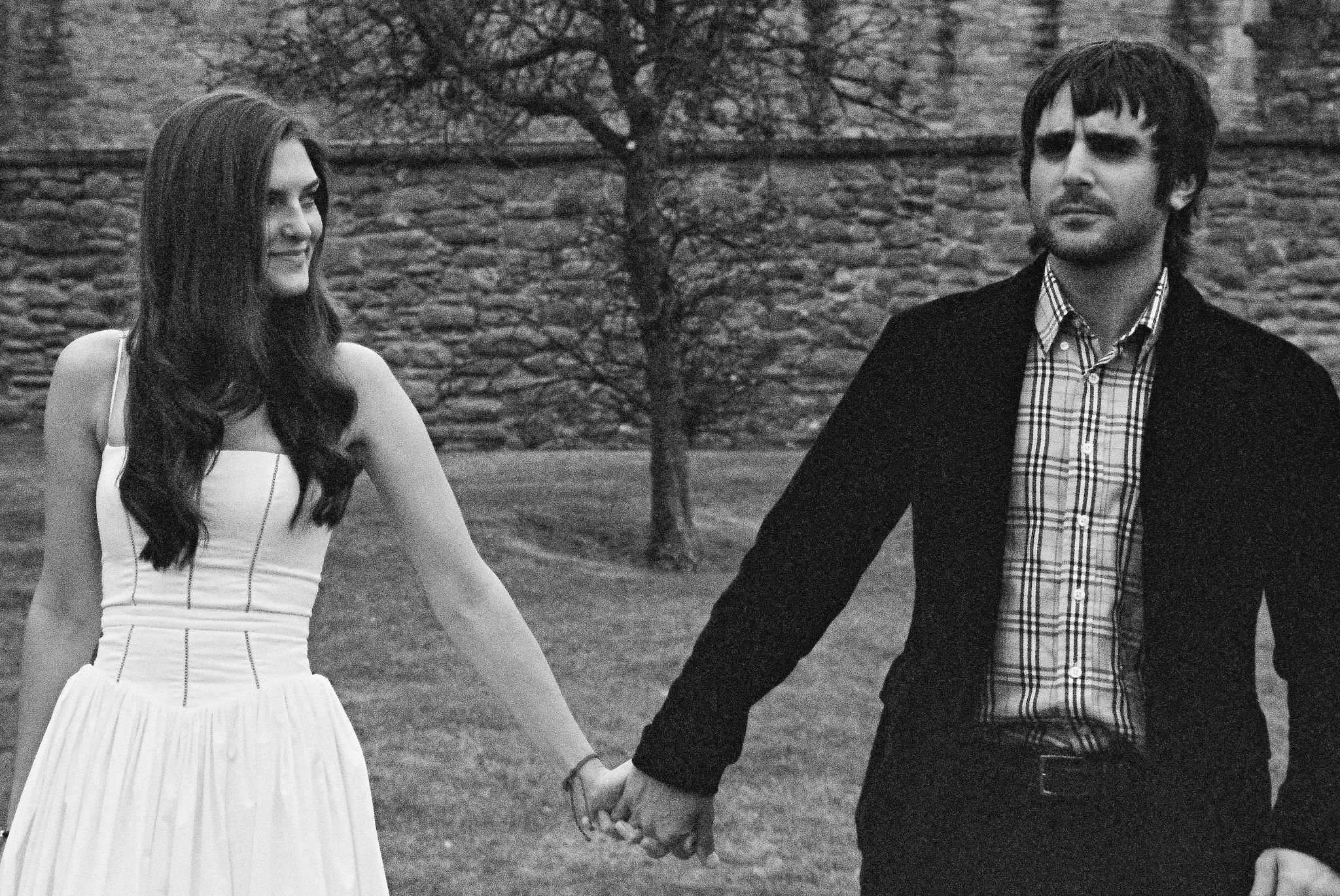 A black and white photo of a young couple holding hands outdoors, with the woman on the left looking at the man, who is on the right looking away. The background features a stone wall and a leafless tree.