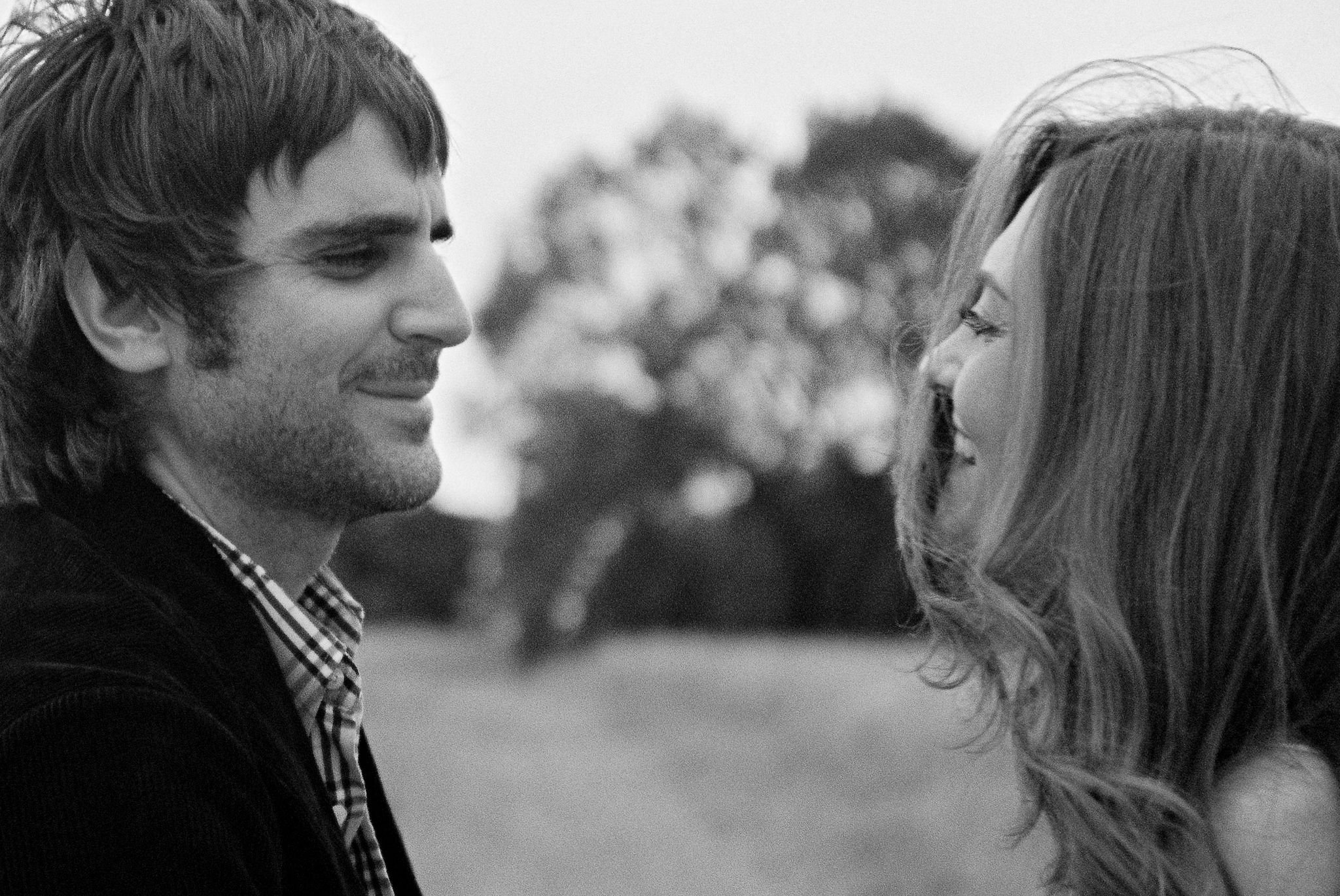 A man and woman face each other outdoors, smiling and looking into each other's eyes, with trees in the background.