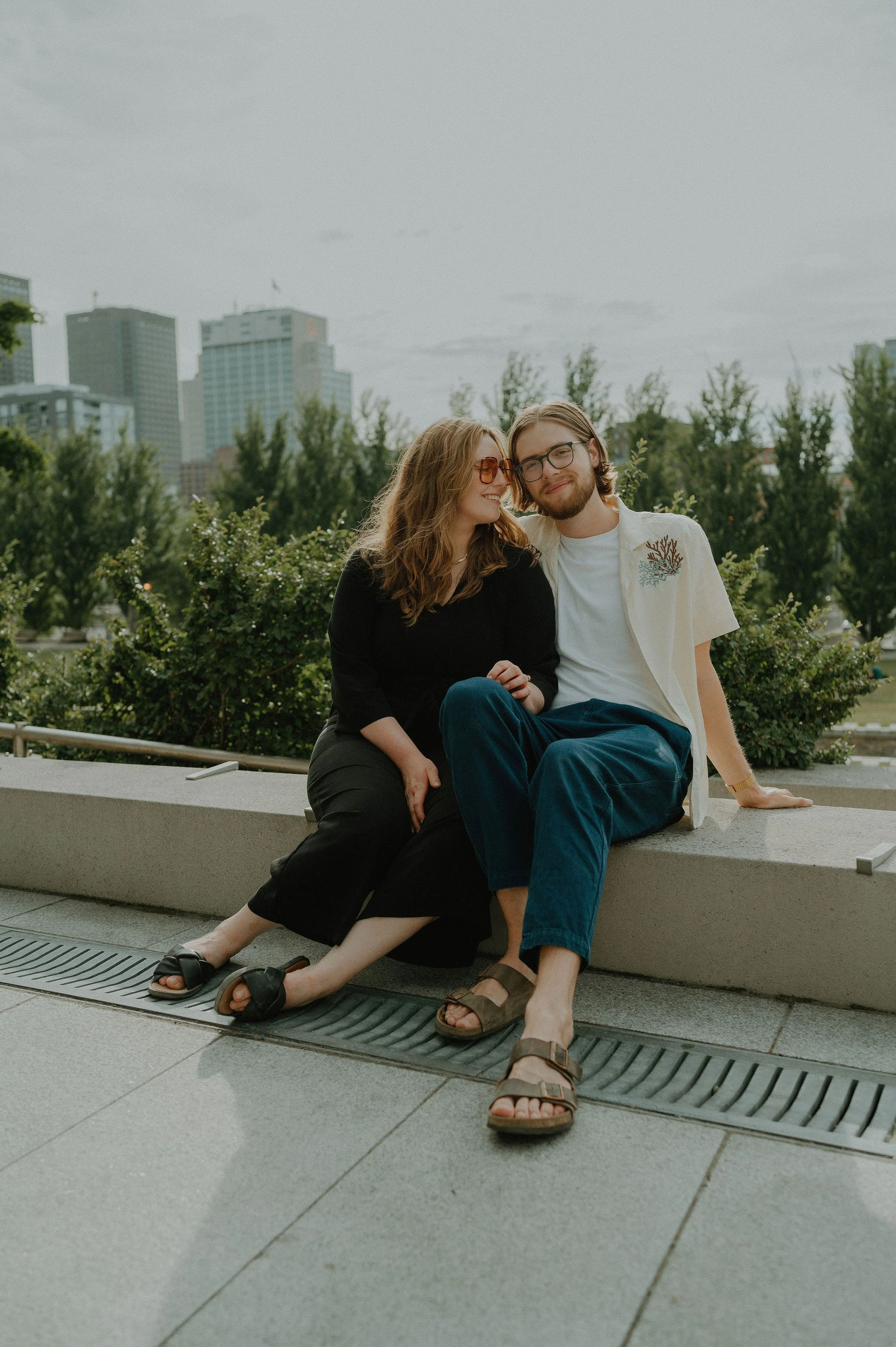 Two friends sitting on a concrete ledge outdoors in an urban park, smiling and enjoying each other's company, with trees and city buildings in the background.