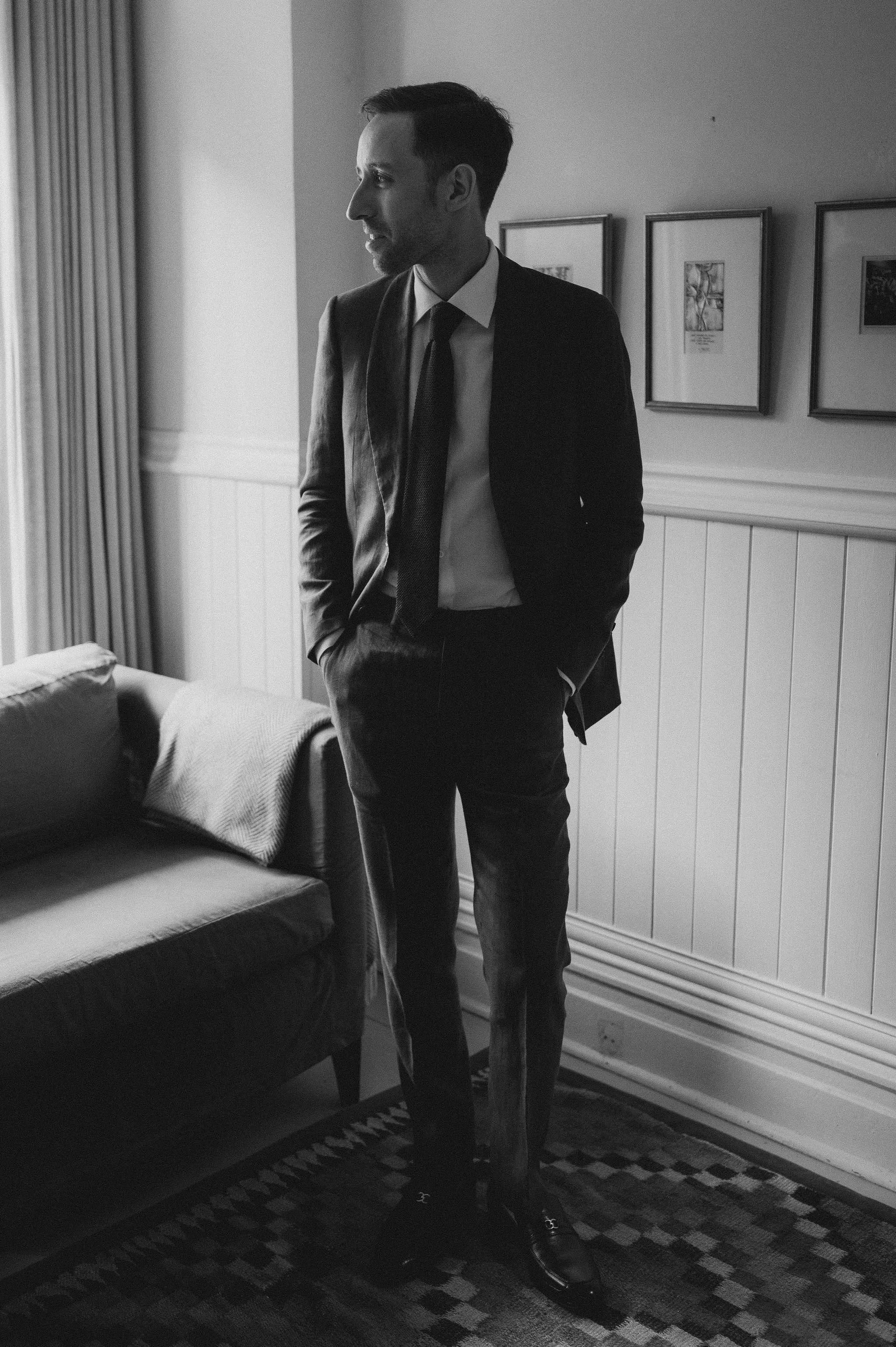 Man in a suit standing by a window with hand in pocket, in a room with framed pictures on the wall.