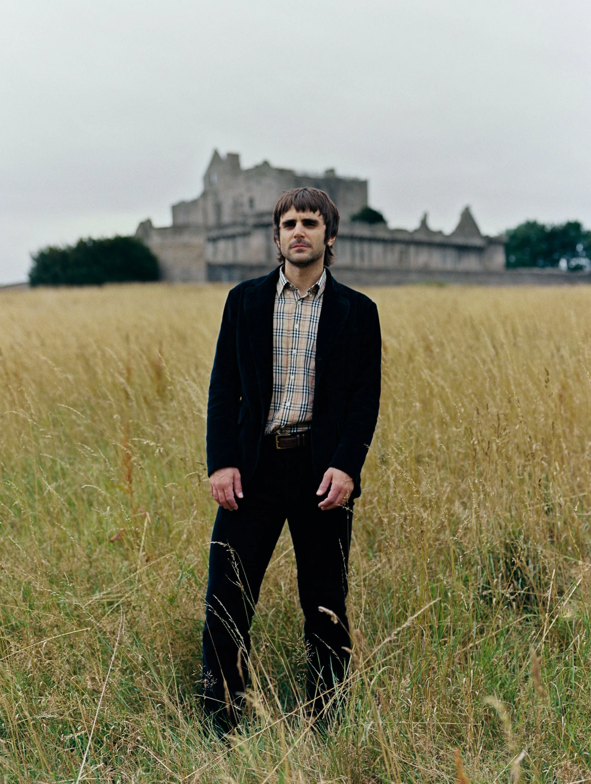 A man standing in a grassy field with a castle in the background on a cloudy day.