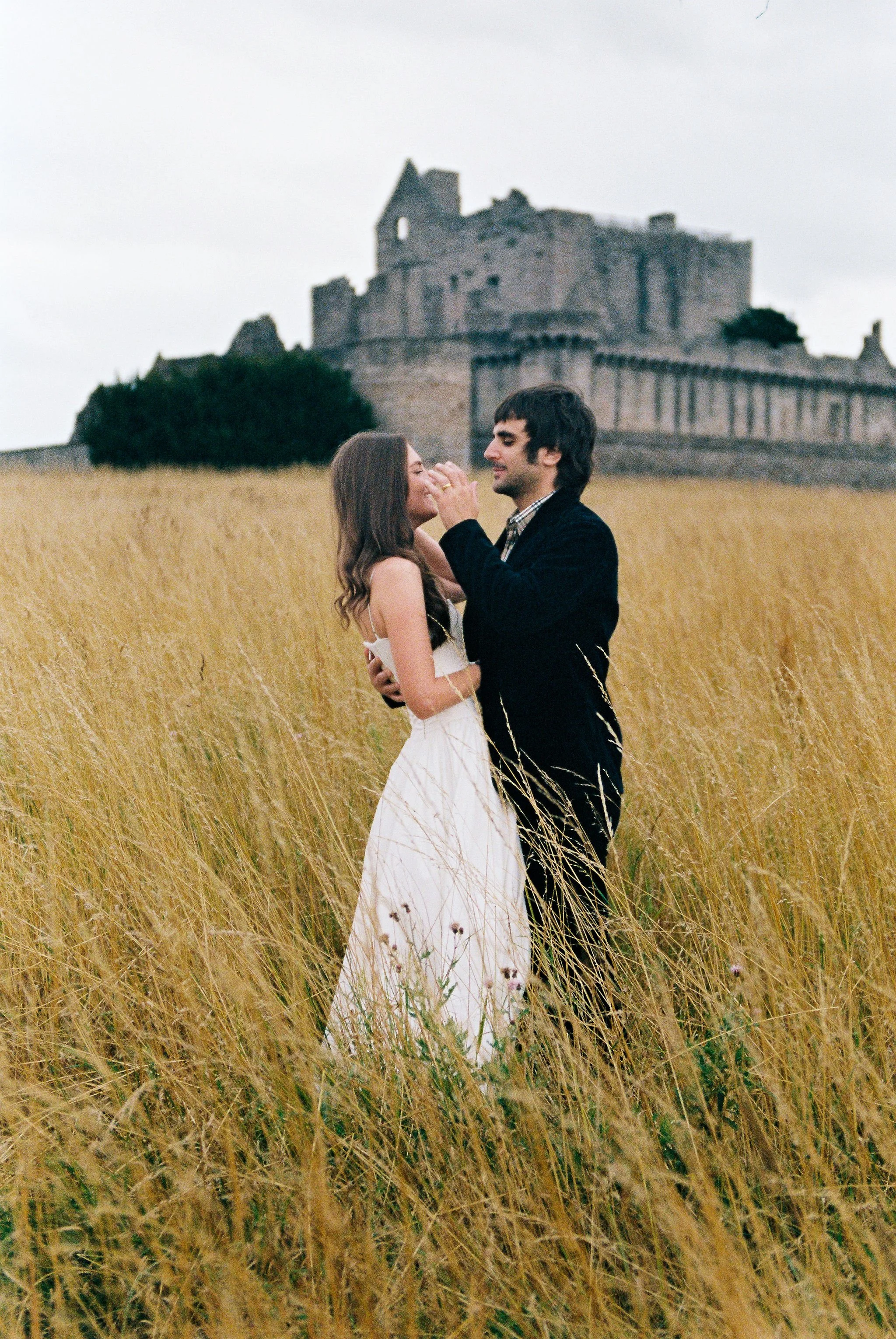 A young couple stands in a grassy field near a castle, sharing an intimate moment during daytime.