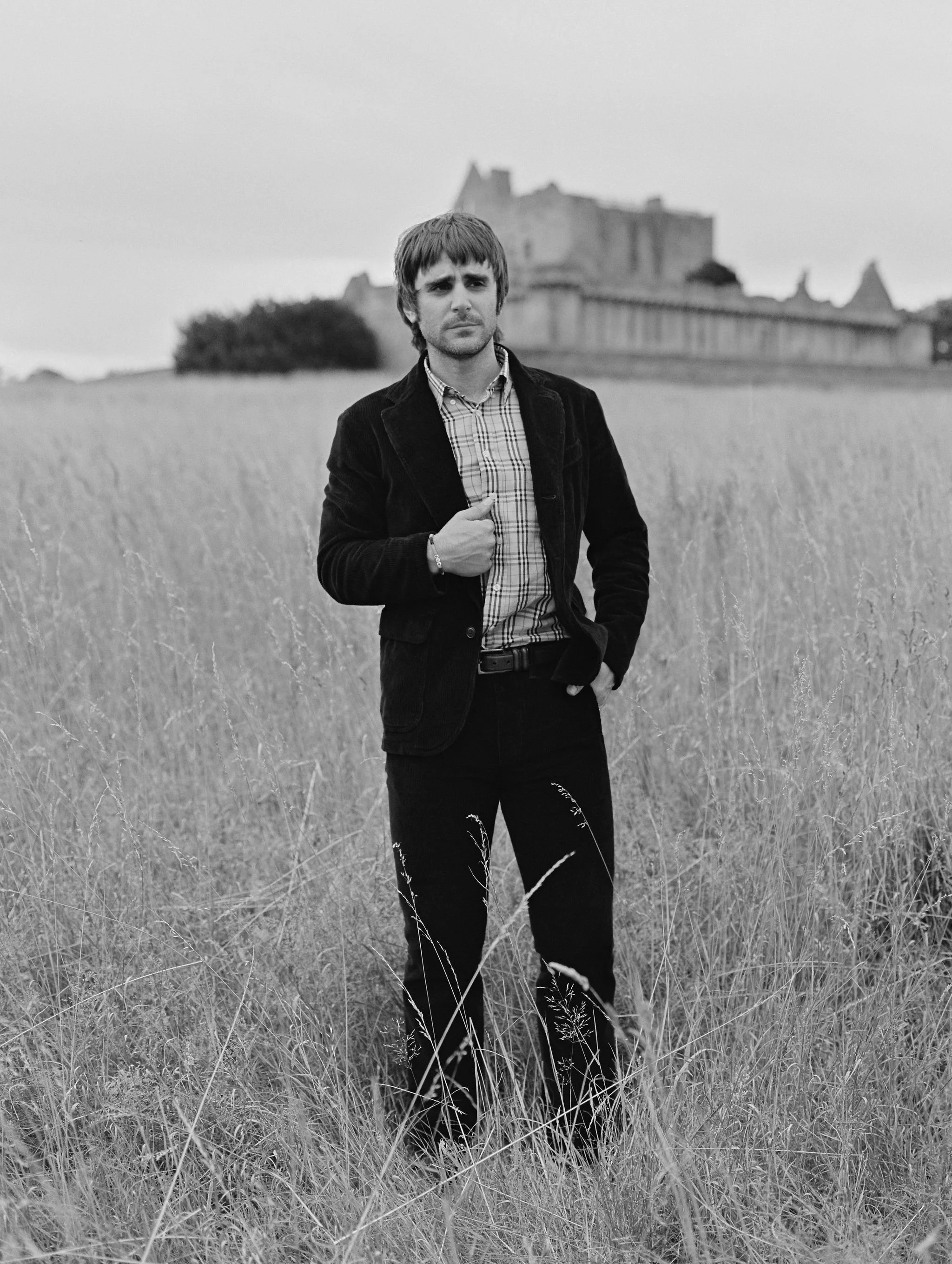A young man standing in a field of tall grass with a castle in the background, black and white photo.