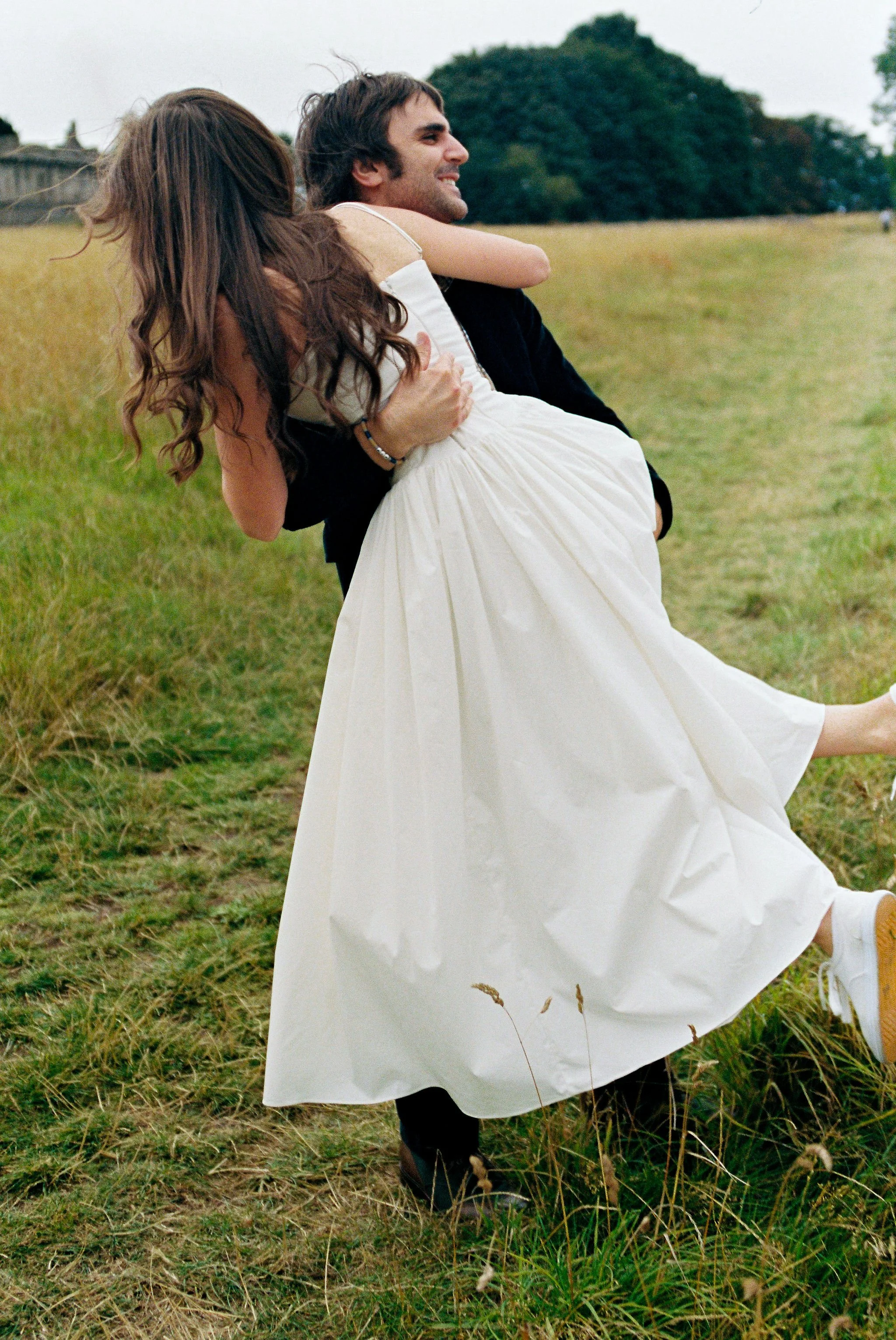 A man in a dark jacket carrying a woman in a white dress across a grassy field.