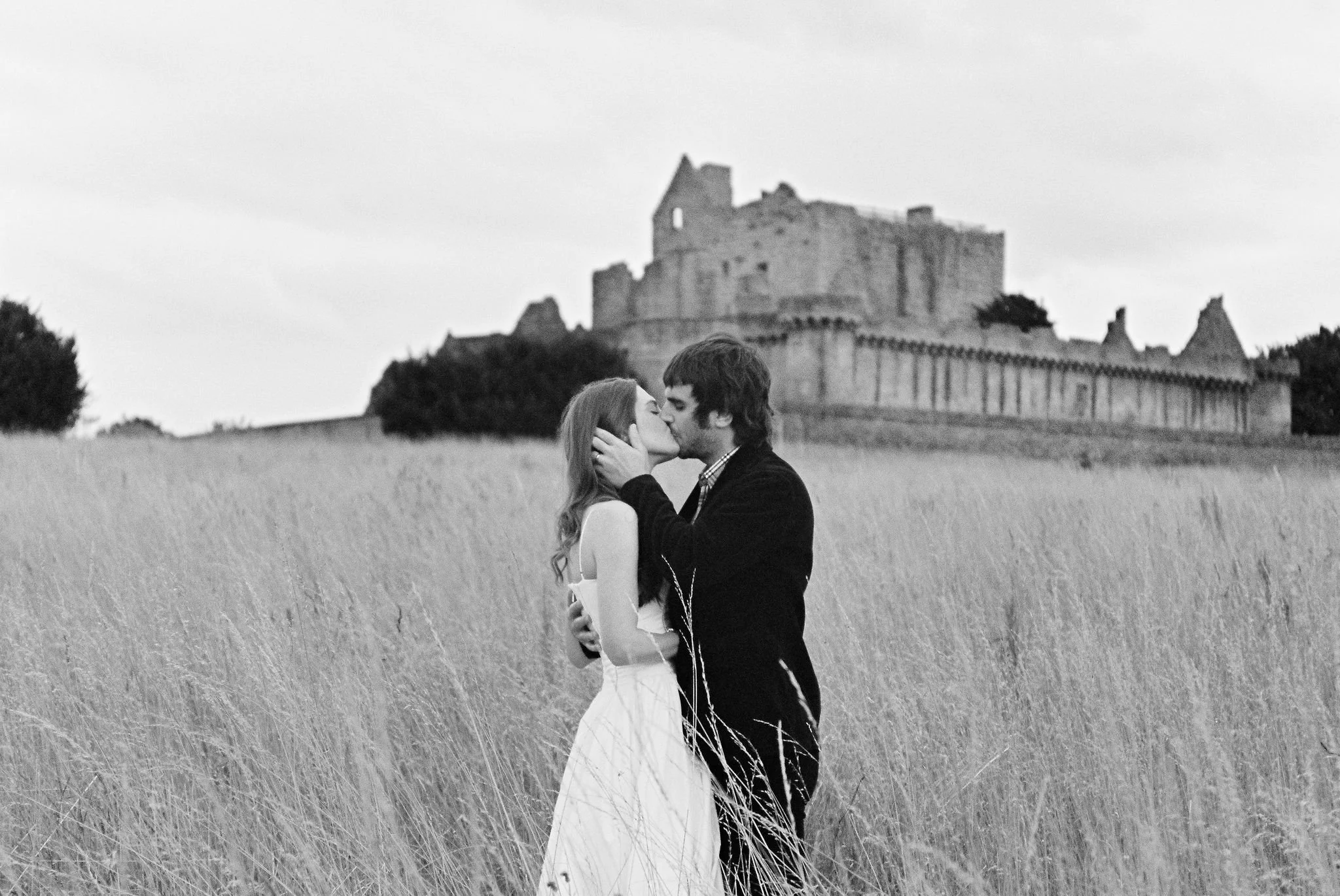 A black and white photo of a couple kissing in a grassy field with a castle in the background.