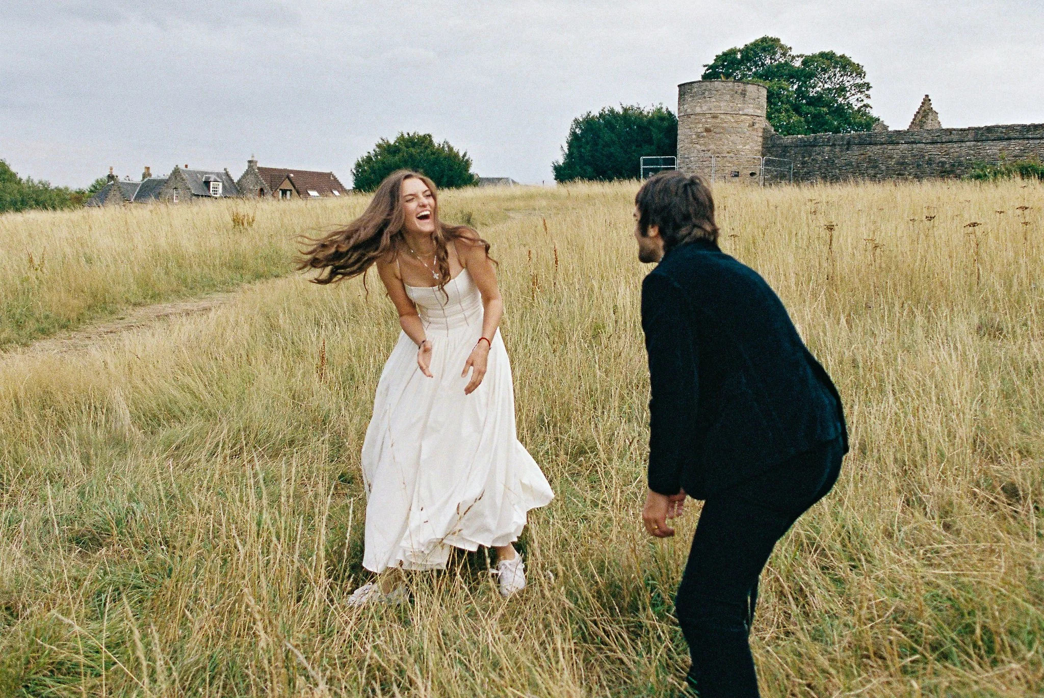 A young woman in a white dress laughing while running through a grassy field, with a young man in dark clothing facing her, in front of a historic stone structure and village houses.
