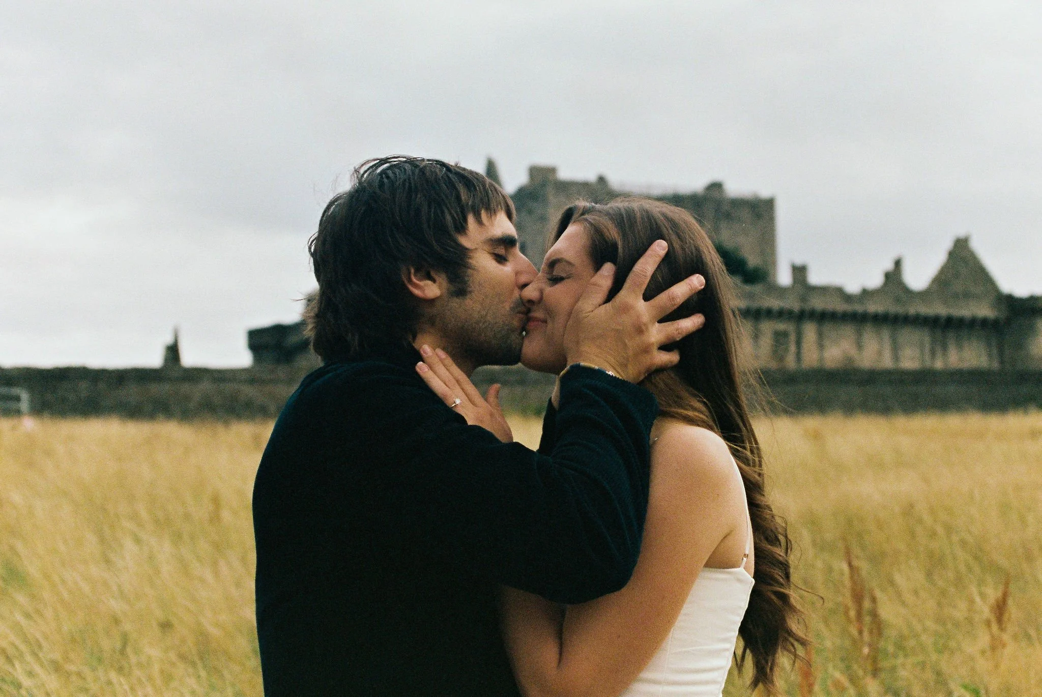 A young couple sharing a kiss in a field with a castle in the background.