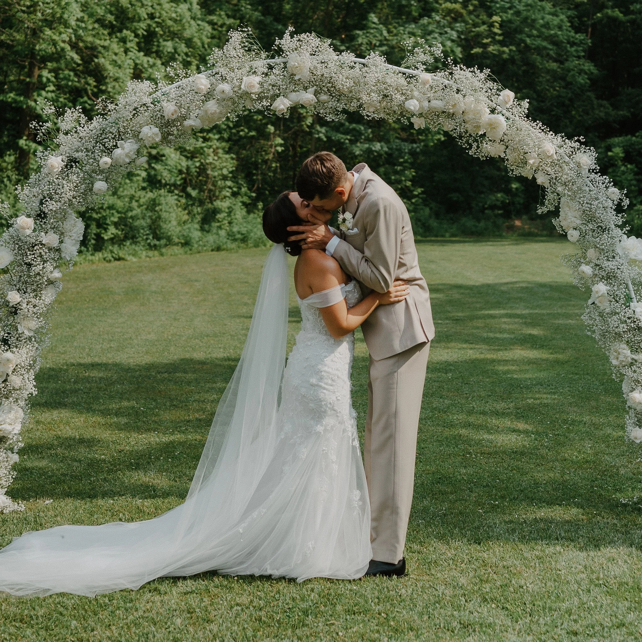 A bride and groom kissing under a floral arch during their outdoor wedding ceremony, with lush green trees in the background.