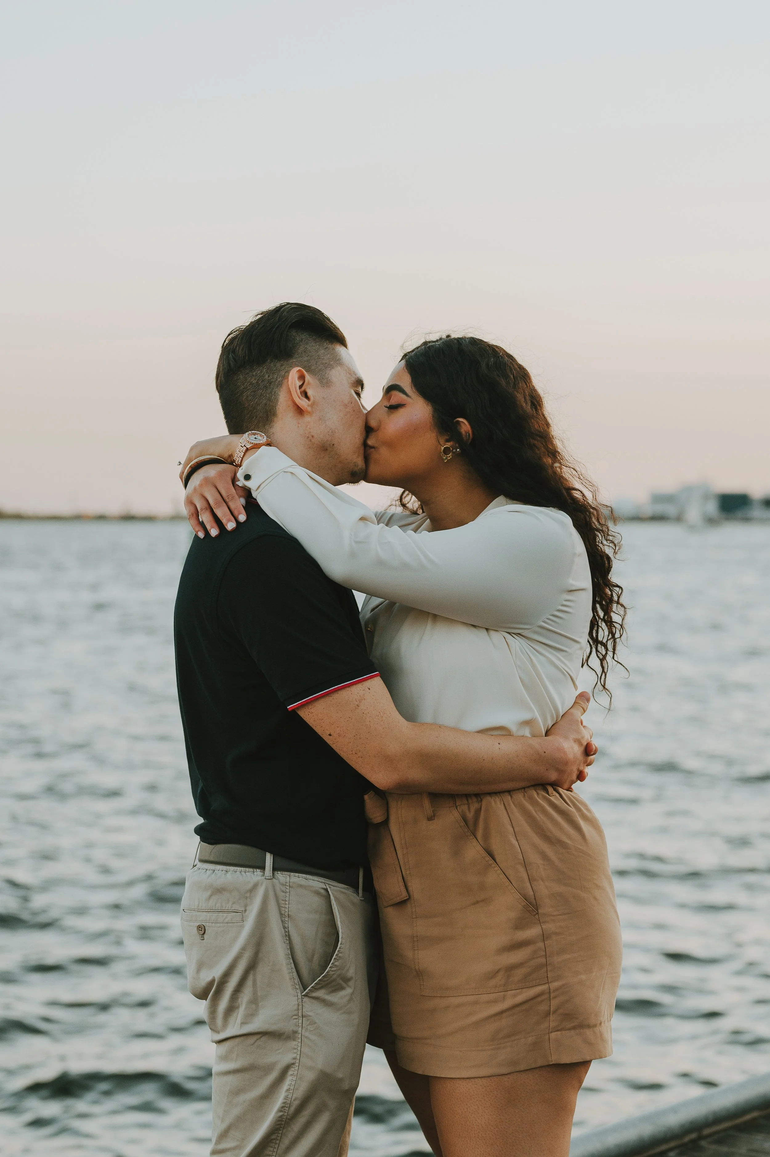 A couple kissing near a body of water at sunset, with city skyline in the background.
