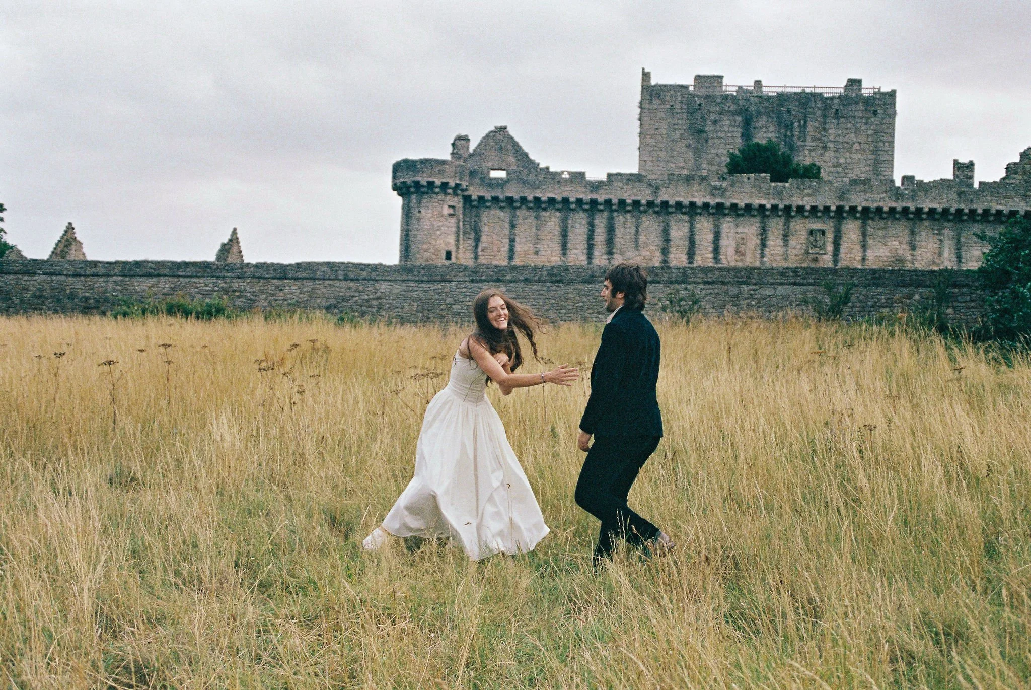 A woman in a white dress and a man in a black suit dancing on a grassy field with a castle in the background.