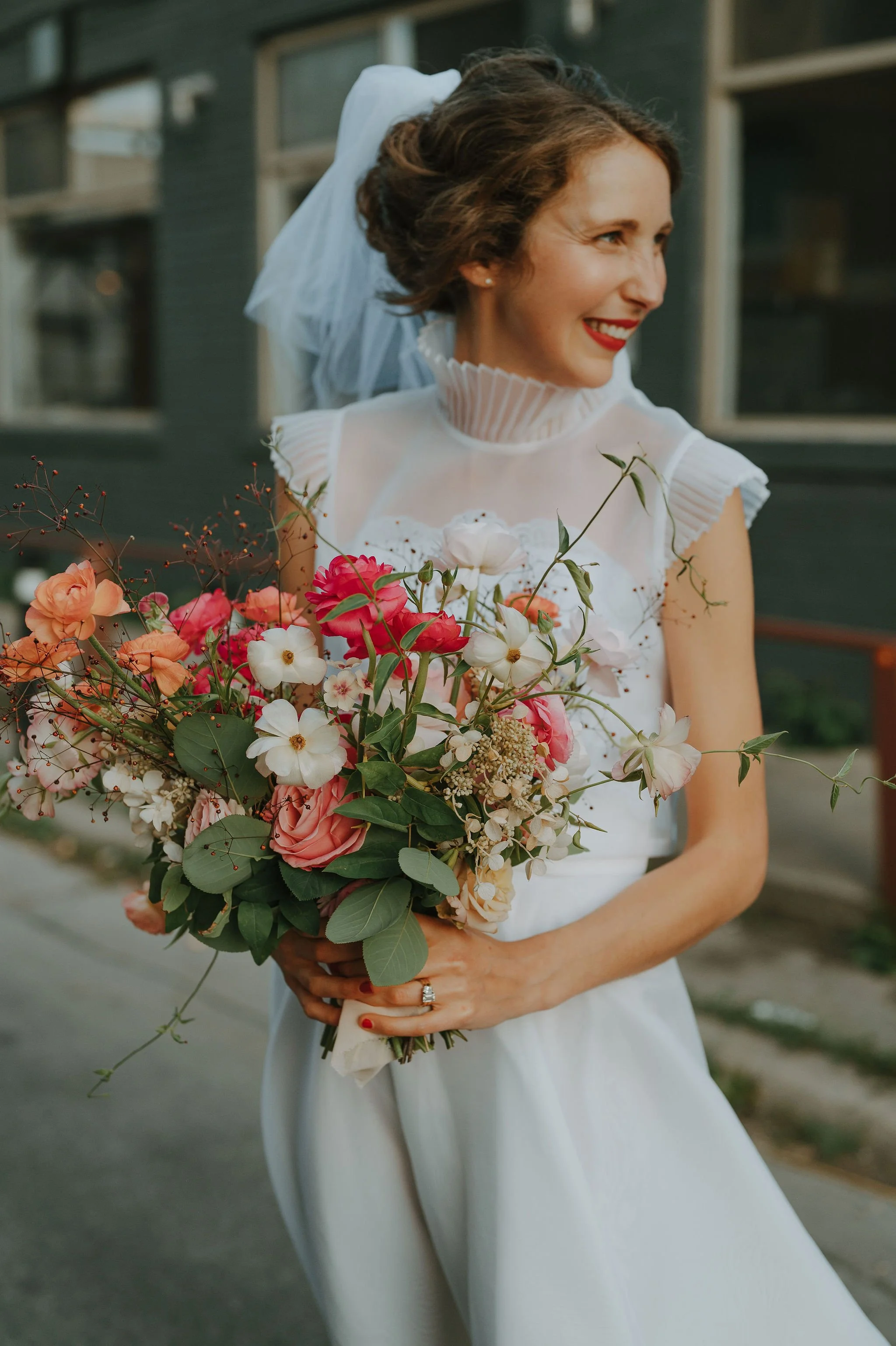 A woman in a white wedding dress holding a colorful bouquet of flowers and smiling outdoors.