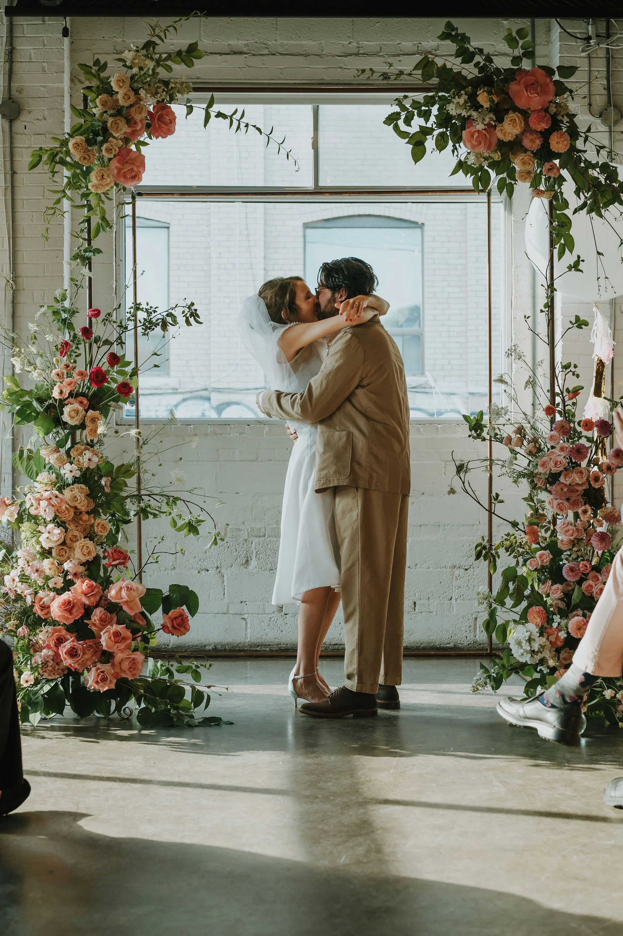A bride and groom share a kiss during their wedding ceremony, framed by a floral arch with pink and white flowers, indoors with natural light coming from a large window.