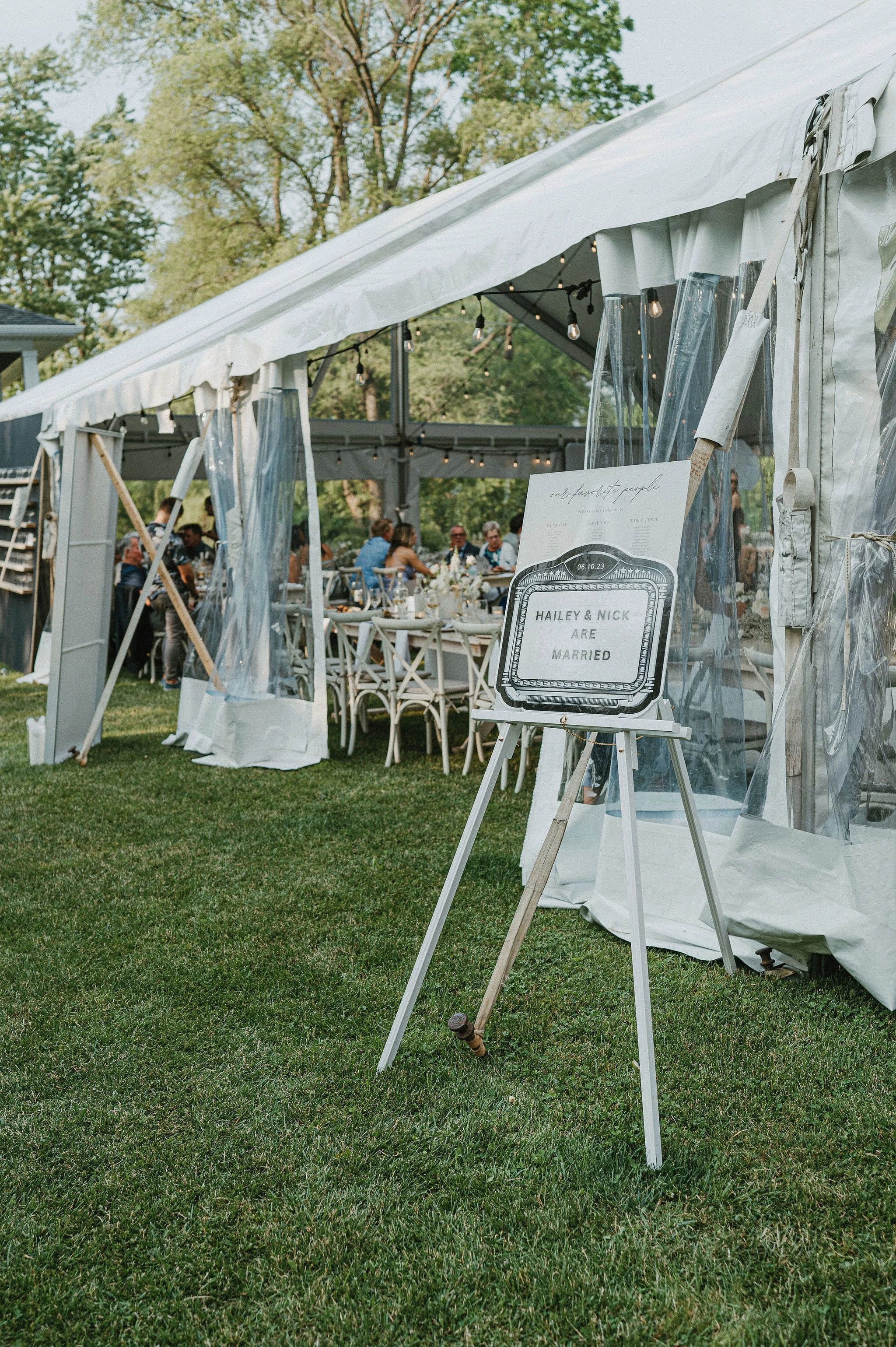 Wedding reception setup under a tent with guests seated at tables, and a sign on an easel reading "Hailey & Nick Are Married."
