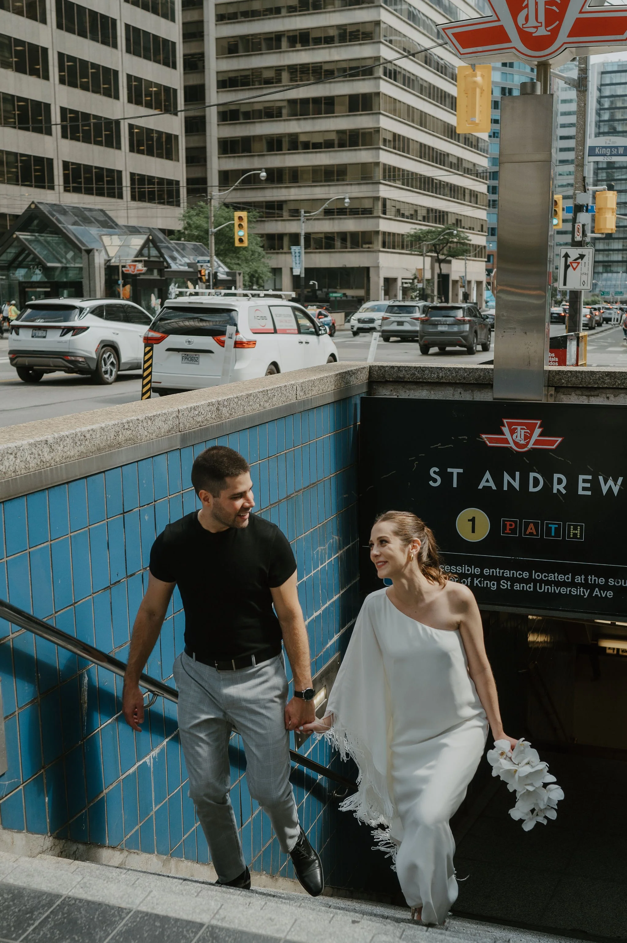 A smiling man and woman holding hands and ascending the stairs at a subway entrance in an urban area. The woman is dressed in a white wedding gown and holding a bouquet. The man is wearing a black t-shirt and gray pants.