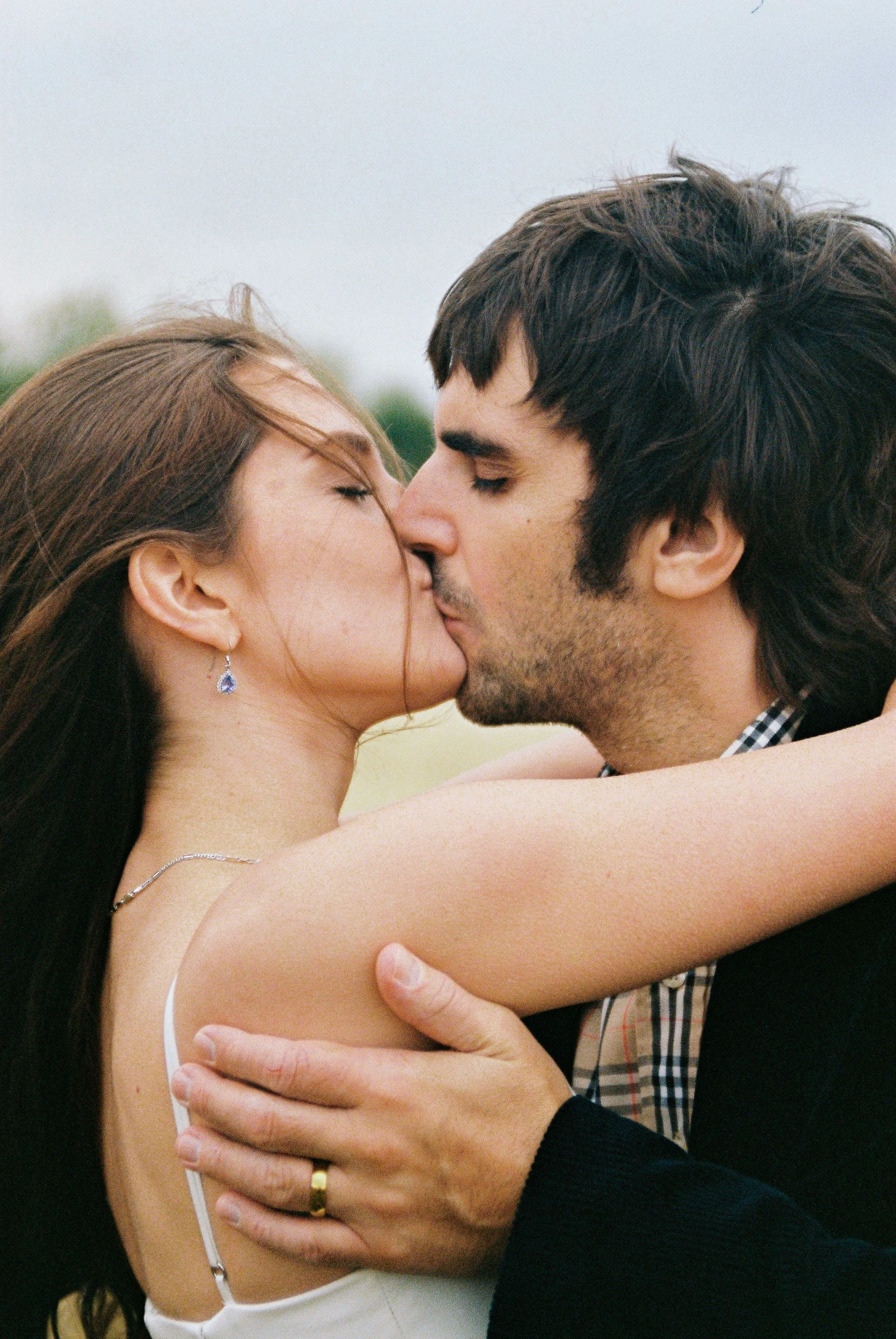 A man and woman kissing outdoors, in an embrace, with the woman’s arm around the man's shoulders.