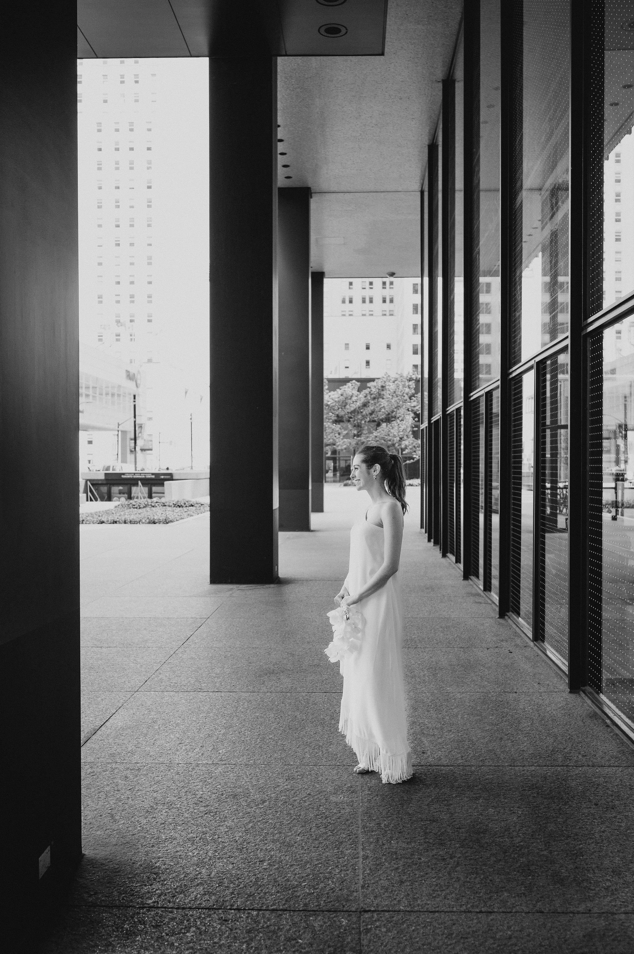 A woman in a white dress standing outside a modern building with glass windows and tall city buildings in the background, holding a bouquet of flowers.