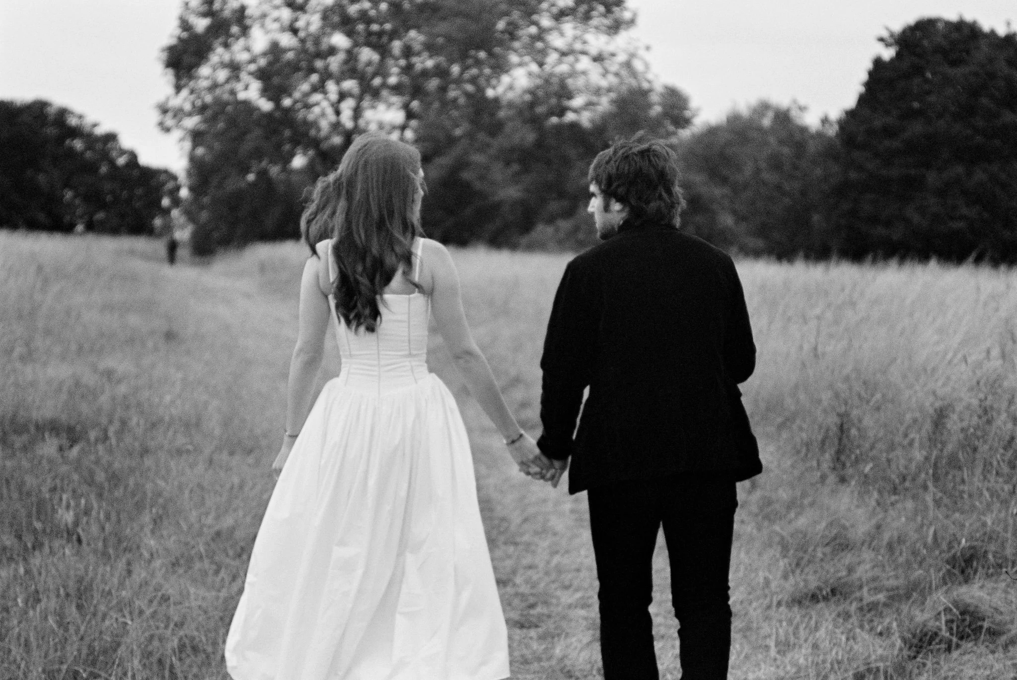 A black-and-white photo of a couple holding hands, walking through a field with trees in the distance, with the woman in a wedding dress and the man in a dark suit.
