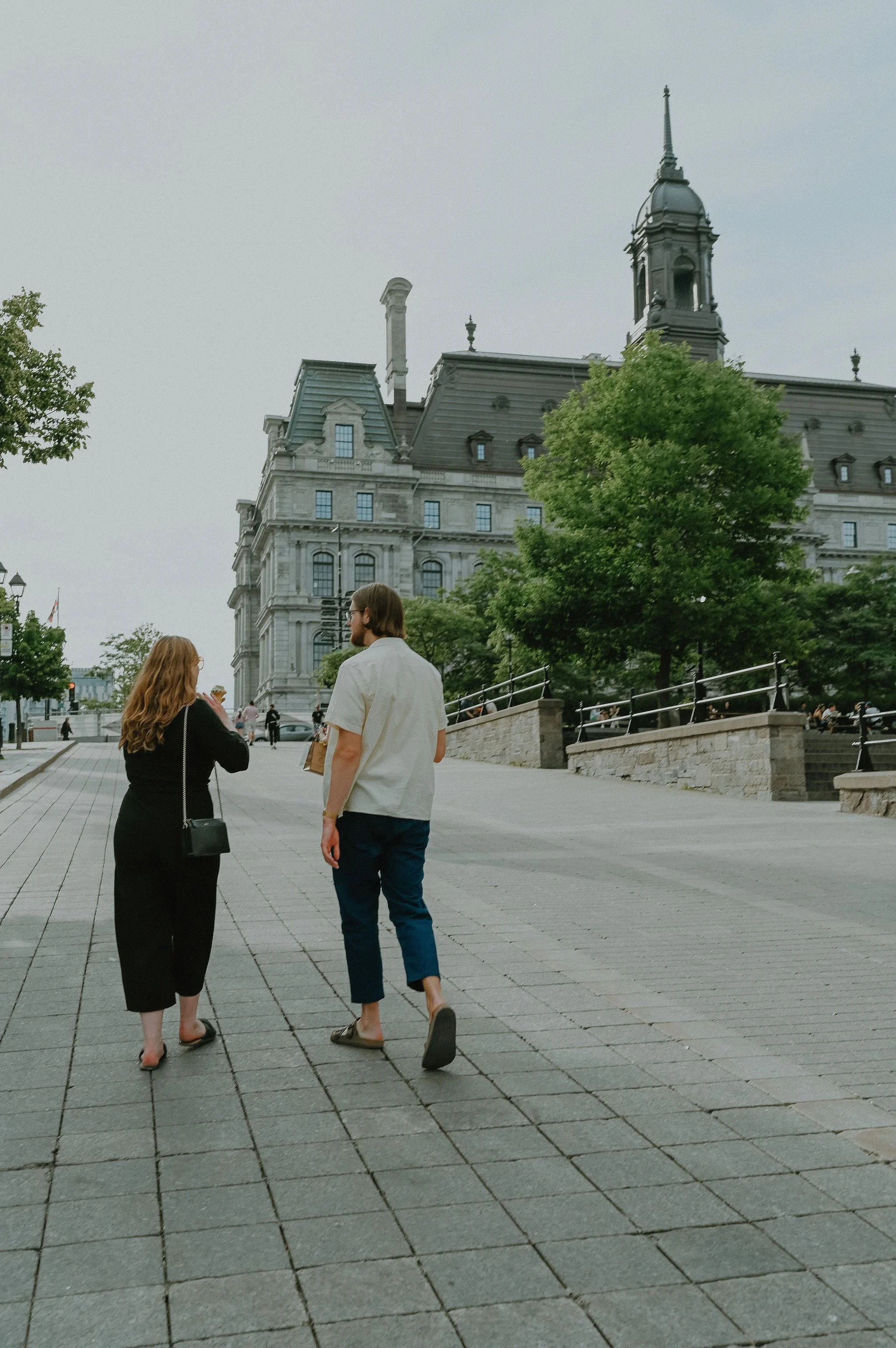 A man and woman walking together on a paved city street near a historic building with a clock tower and trees.