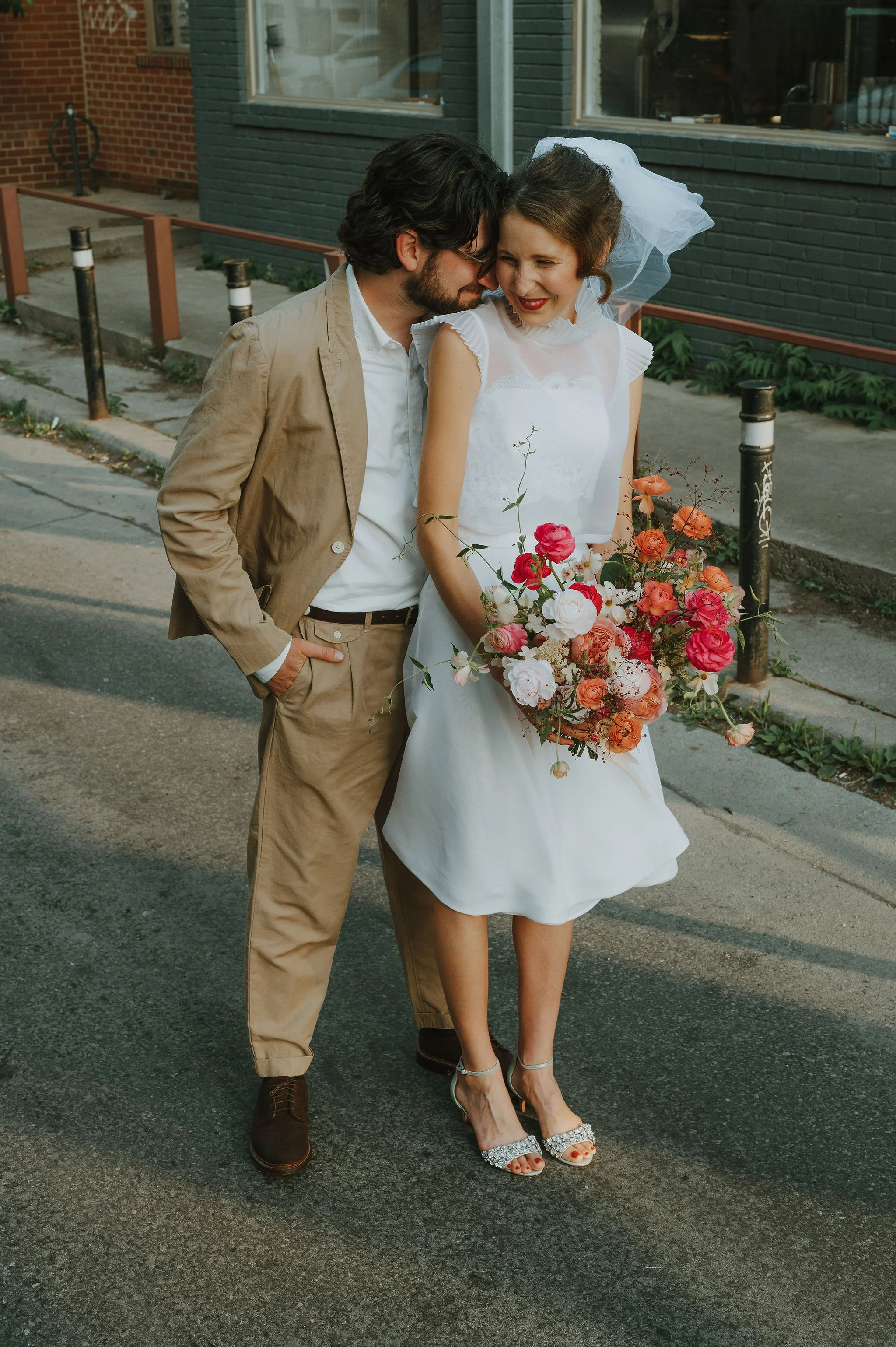 A couple dressed in wedding attire standing on a street, with the woman holding a large bouquet of flowers. They appear to be sharing a tender moment.