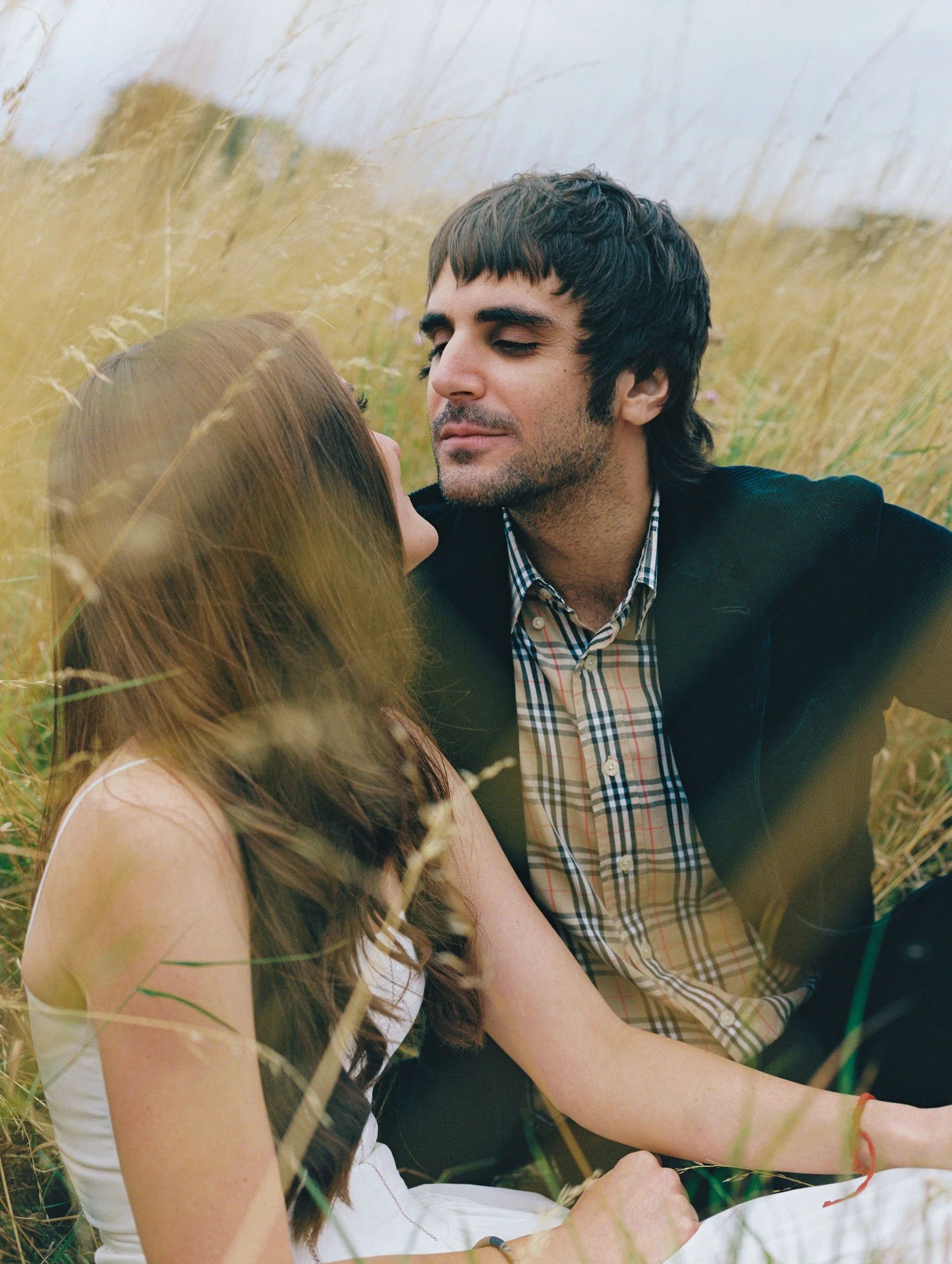 A man and woman sitting in a grassy field, facing each other closely with the man gently leaning in, surrounded by tall grass and a cloudy sky.
