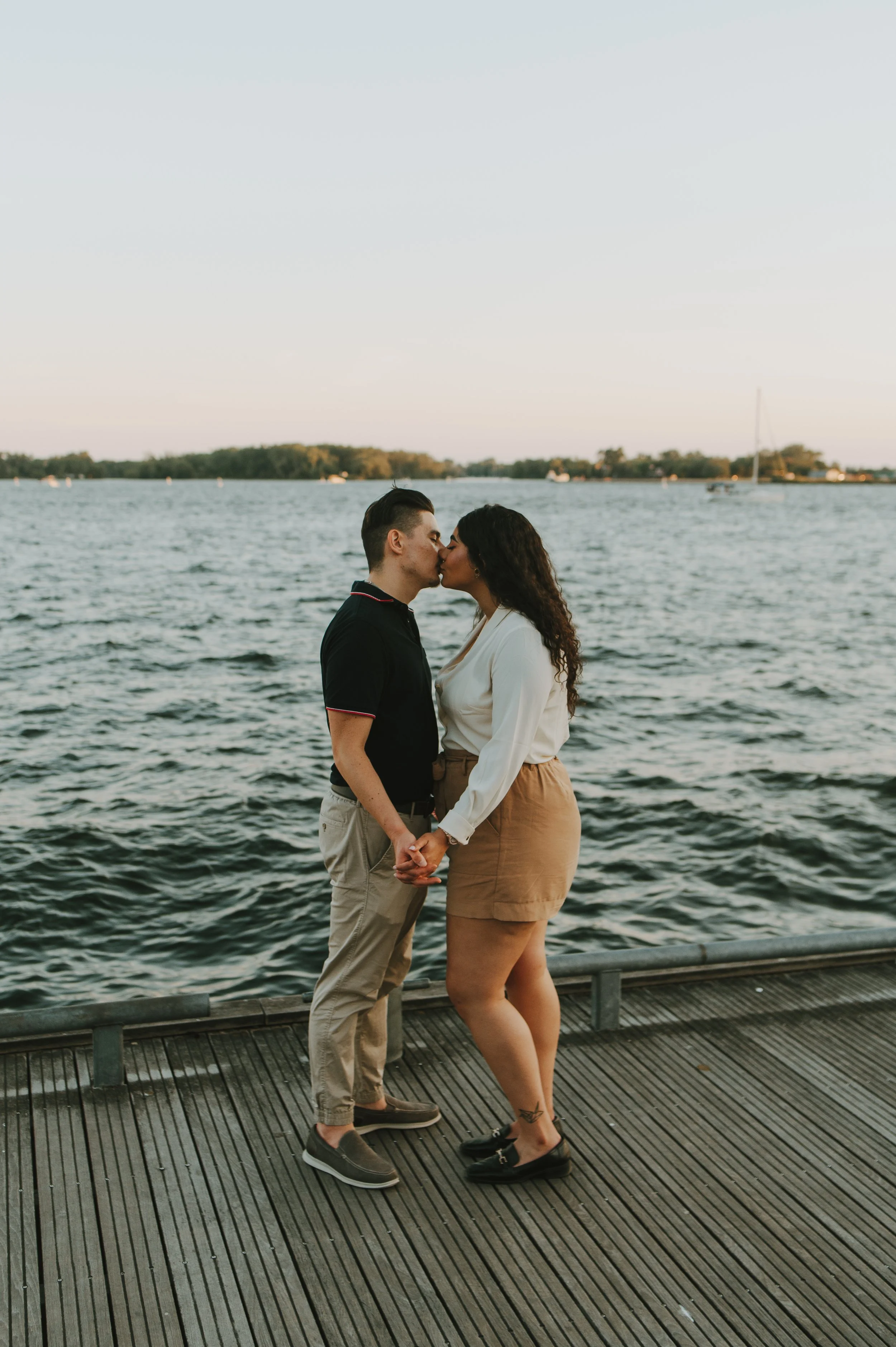 A couple kissing by a body of water, holding hands, standing on a wooden dock during sunset.
