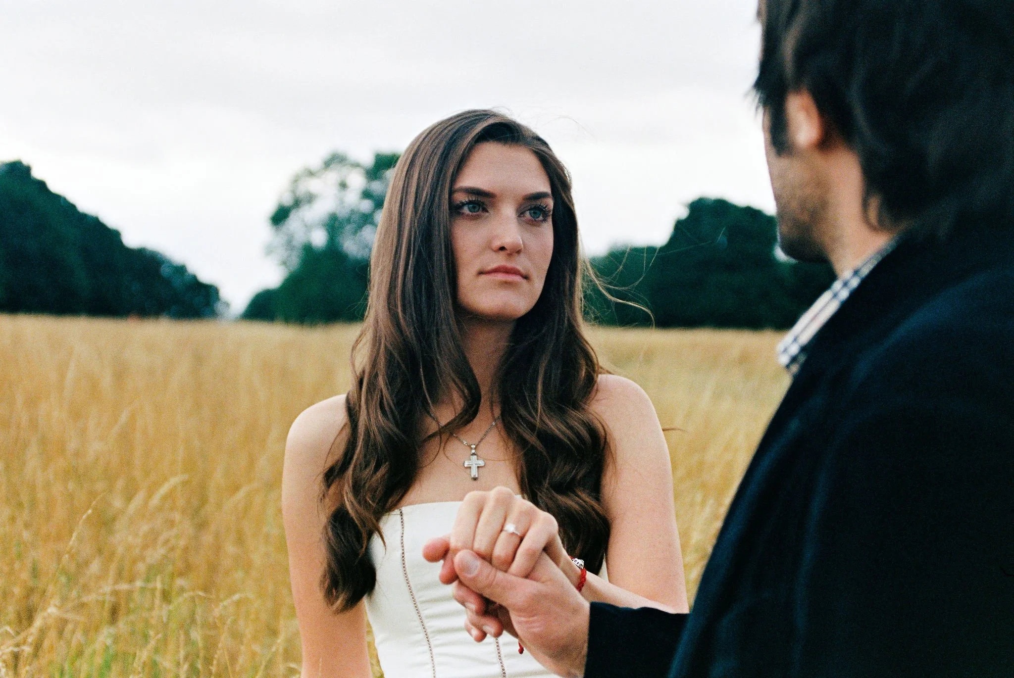 A woman with long, wavy brown hair and a cross necklace holding hands with a man in a black jacket in a wheat field.