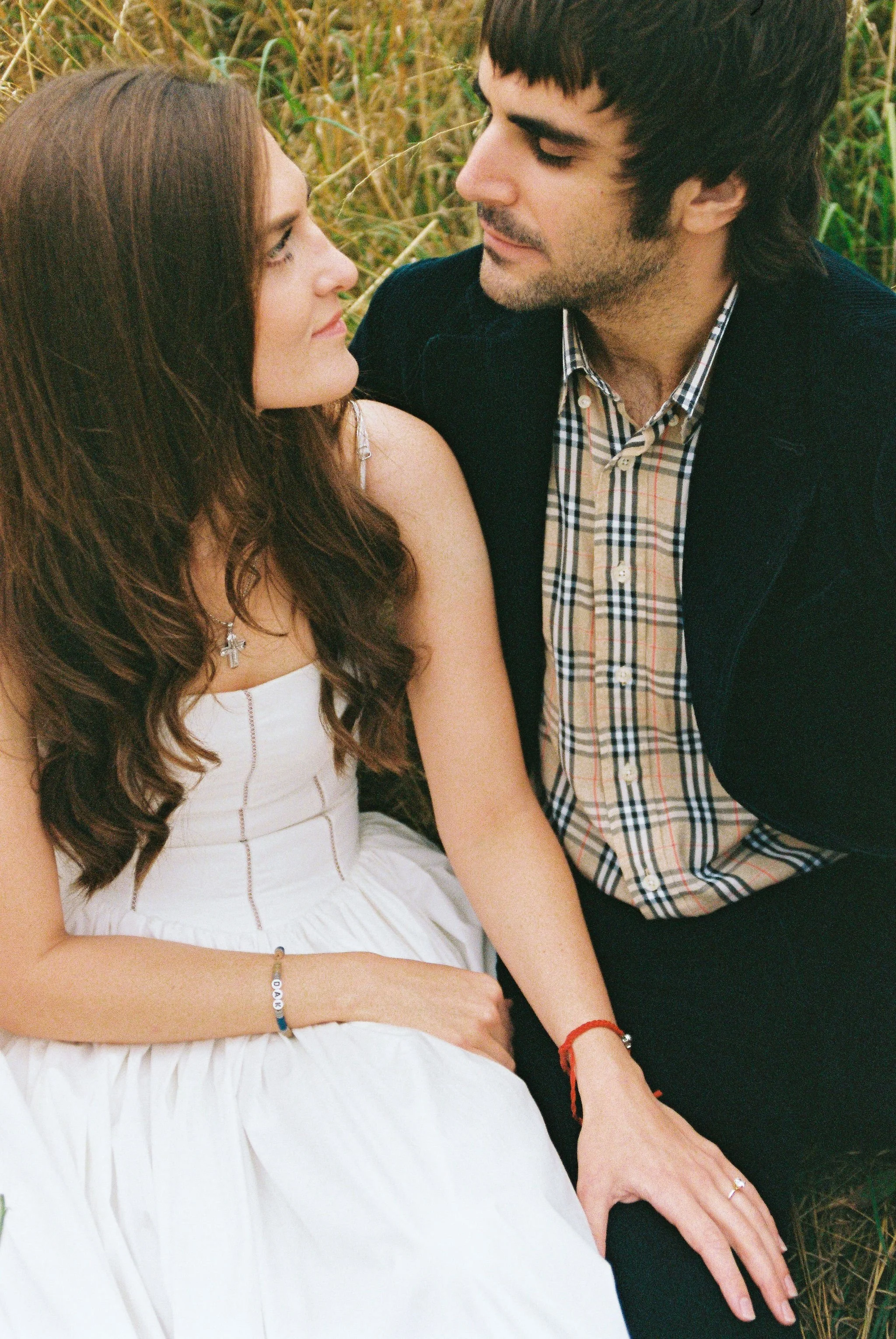 A man and woman sitting close together in a grassy field, looking into each other's eyes.