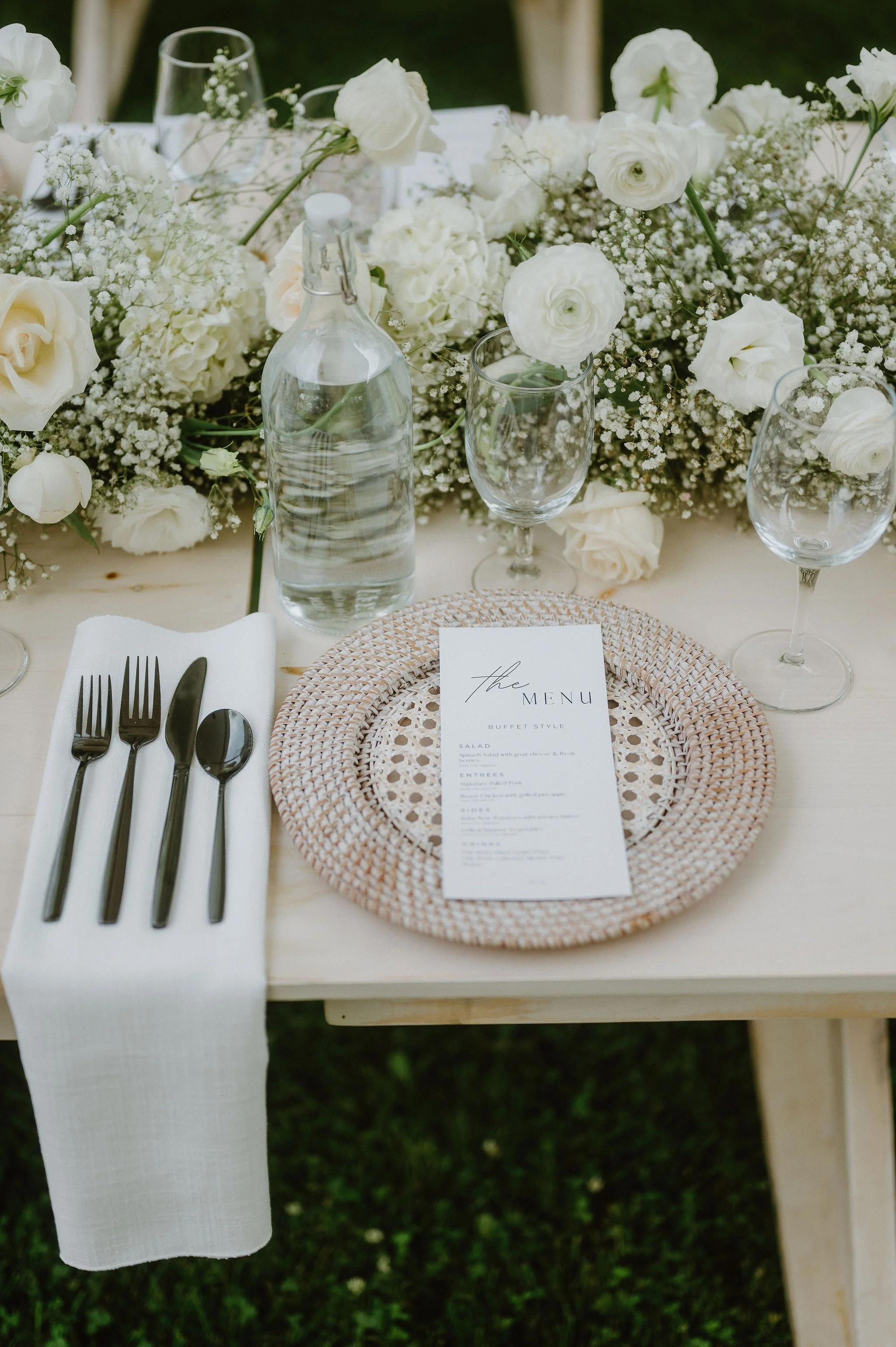 Elegant outdoor table setting with white flowers, a menu, water bottle, and wine glasses arranged on a light-colored wooden table.