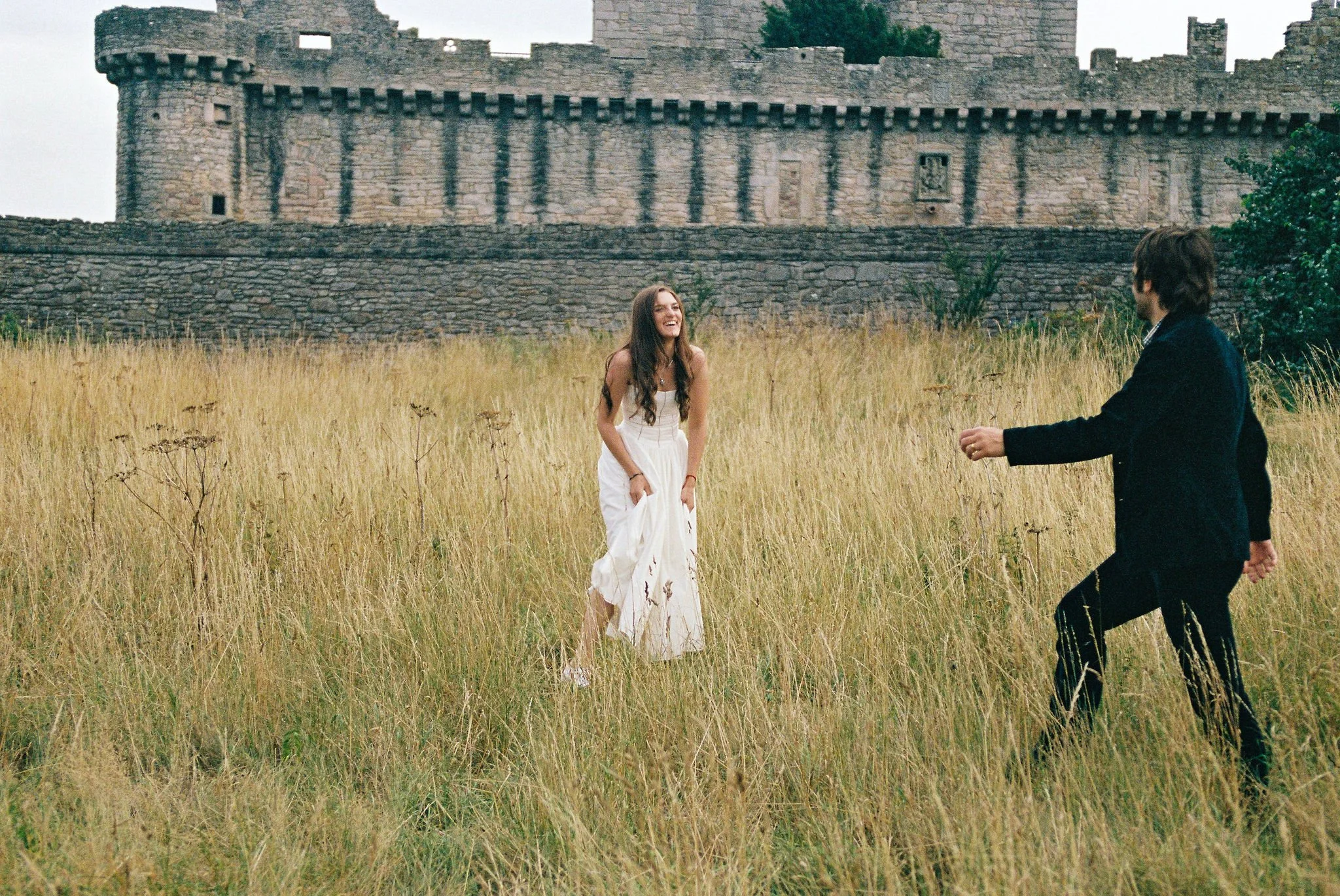 A woman in a white dress laughing and a man in a dark suit walking through a field of tall grass in front of an old stone castle.