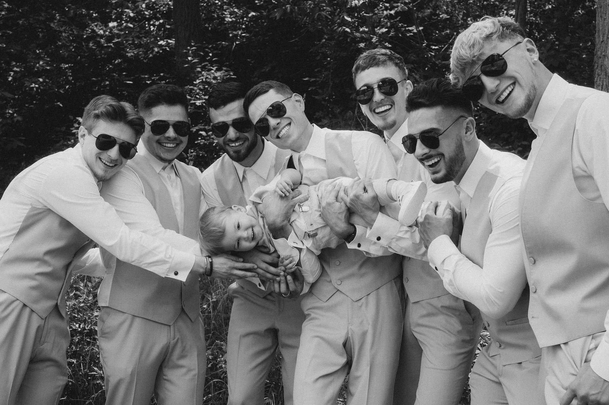 A group of men and a young girl, all smiling and wearing sunglasses, are outside in a wooded area. They are lifting a young girl in the air while posing for the photo.