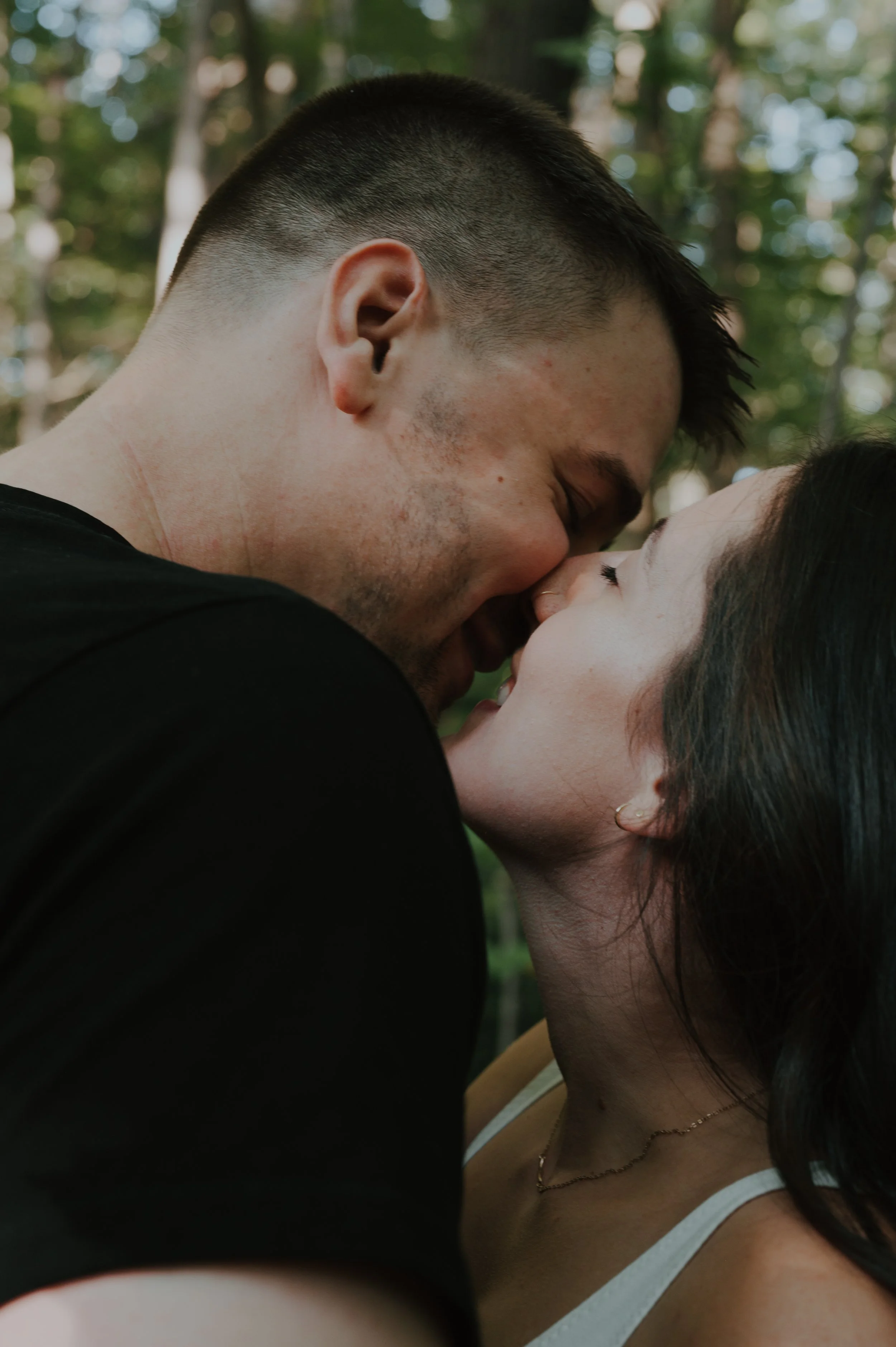 A couple is about to kiss outdoors in a forested area, with the man and woman close together, eyes closed, smiling, and touching noses.