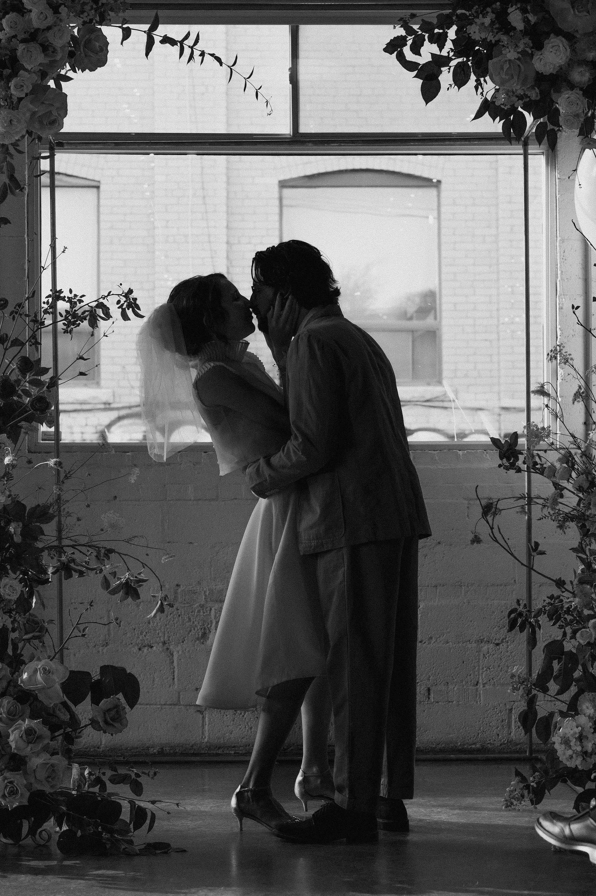 Black and white photo of a couple kissing, with the woman's hand on the man's face, standing in front of a window with brick building outside, decorated with floral arrangements.
